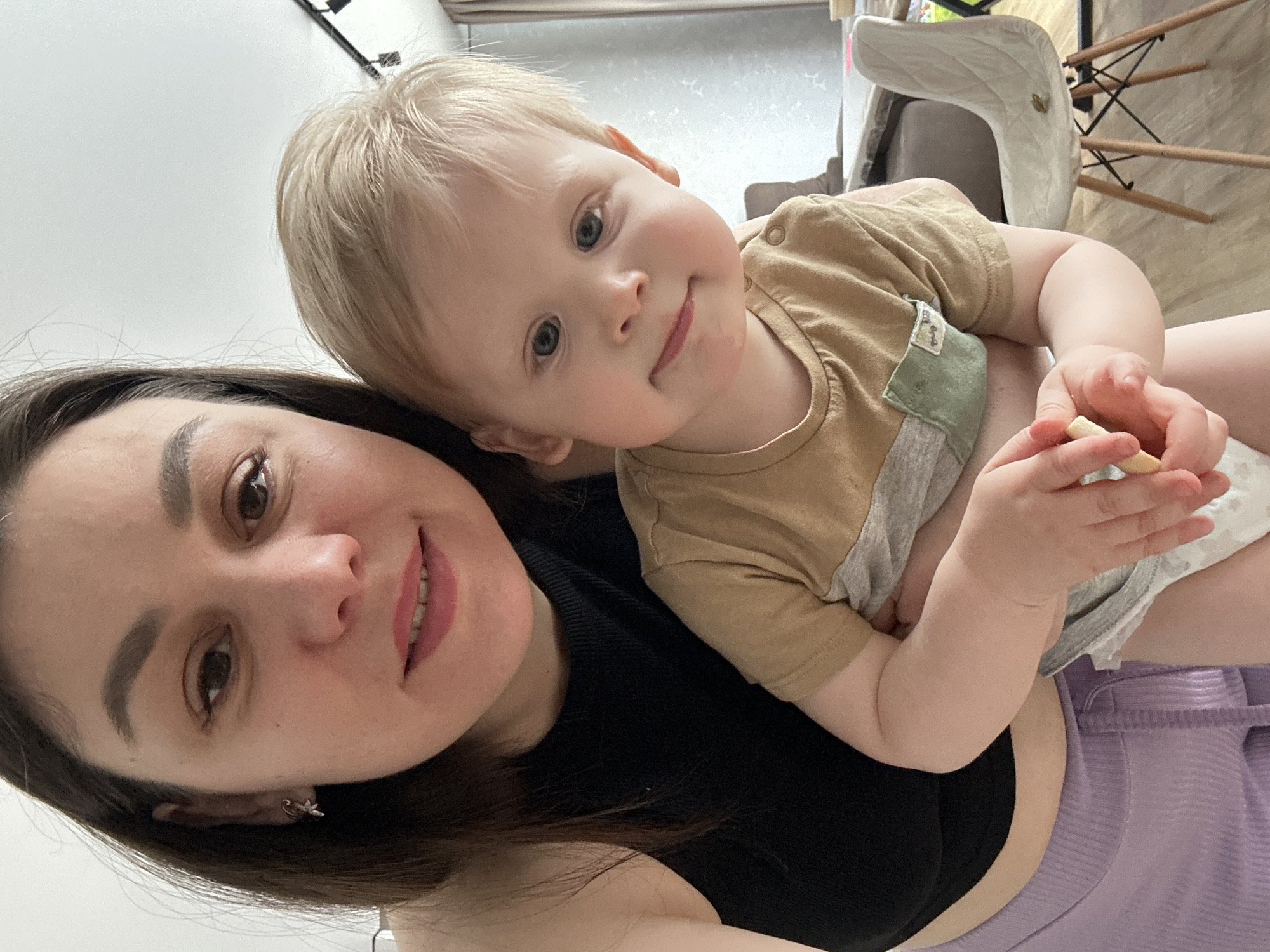 An indoor selfie, taken in Chaikovsky, Russia, features a woman and a young child. The woman, positioned in the foreground, has dark hair and brown eyes, and is looking at the camera with a gentle smile. She is wearing a black top. A blonde-haired, blue-eyed child, appearing to be a toddler, rests their head on the woman's shoulder, also looking at the camera with a soft smile. The child wears a two-tone t-shirt, tan and grey, over a white diaper, and holds a light-colored snack stick in their hands.
The setting appears to be a modern home interior. In the background, parts of a contemporary chair with a light-colored seat and wooden legs are visible, along with light-colored flooring and overhead light fixtures. The woman is also partially seen wearing light purple, ribbed bottoms. The lighting is bright and even, suggesting it is daytime. Both individuals display expressions of happiness and contentment, highlighting a tender moment between them. No visible text is present in the image.