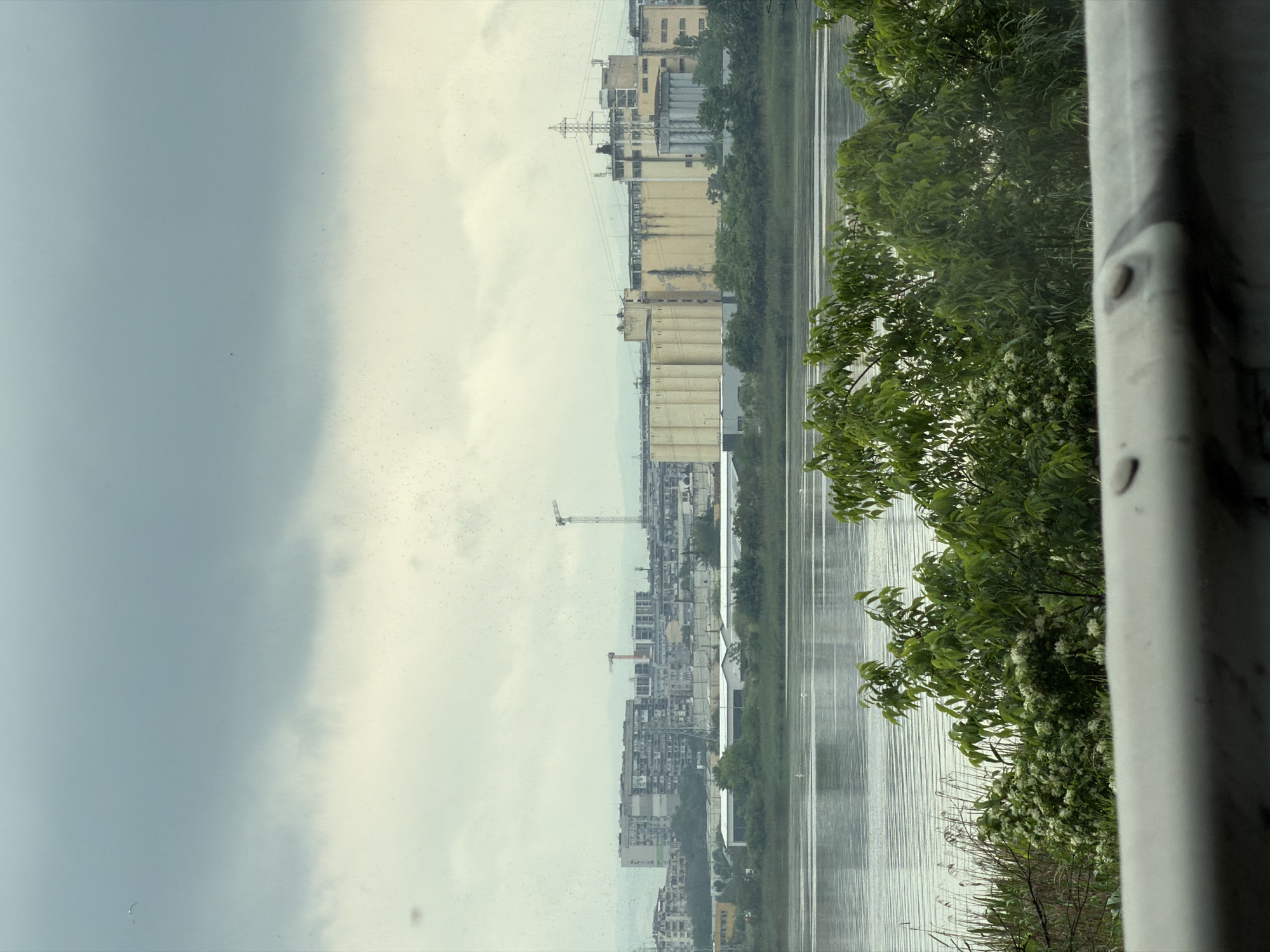 The image depicts a waterfront scene in Burgas, Bulgaria, taken from an elevated perspective, likely from a building or bridge. The foreground shows part of a gray concrete railing. The middle ground features a body of water, possibly the Black Sea or a lake, reflecting the overcast sky. Adjacent to the water, there are trees and vegetation. In the background, there is a cityscape with buildings of varying heights and architectural styles, including grain silos, newer high-rise structures, and cranes, which suggest ongoing construction. The sky is overcast and gray.