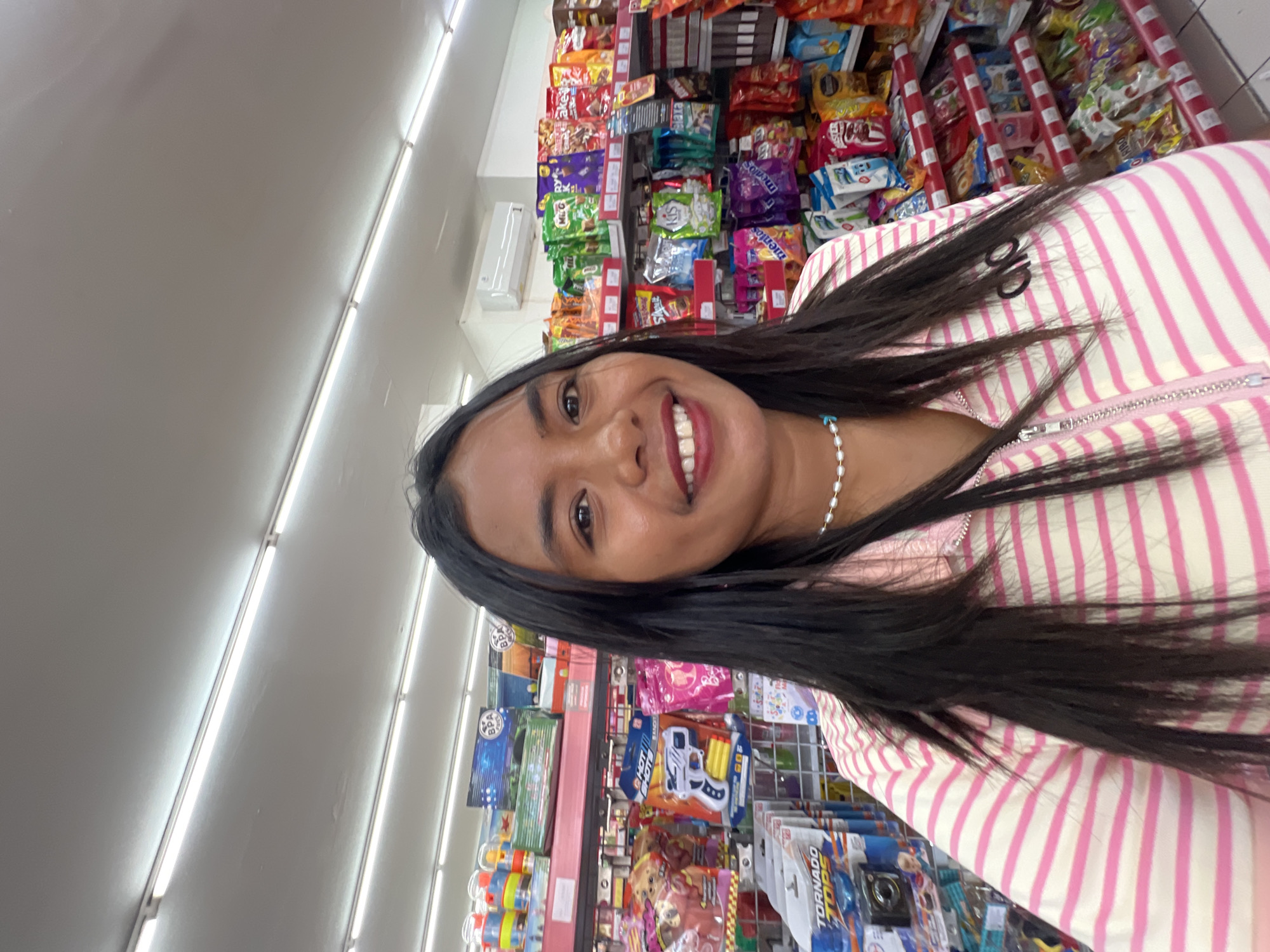A young Indonesian woman with dark hair and a pearl necklace smiles as she takes a selfie in a well-lit convenience store. She is wearing a pink and white striped zip-up jacket with the word "alo" printed in black on the right side of her chest. The background shows shelves stocked with various snacks, toys, and household items. The store appears to be a modern retail space with fluorescent lighting. Based on the context, the location is likely a minimart in Kuta, Indonesia, a popular tourist area known for its many convenience stores.