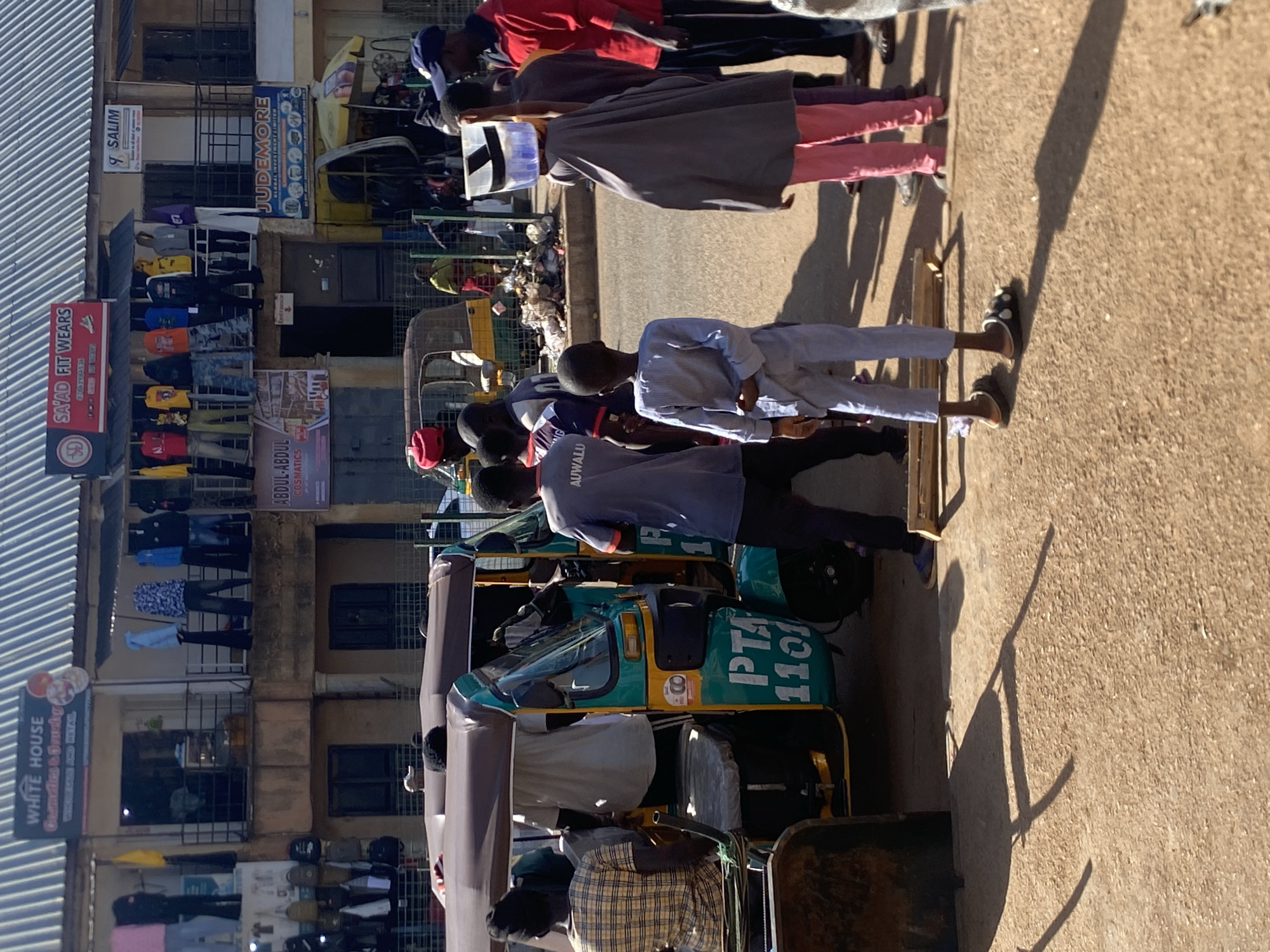 The image depicts a street scene in Bukuru, Nigeria, with a focus on a three-wheeled vehicle, possibly a tuk-tuk, parked on a dirt road. The vehicle is teal and yellow, with white lettering on its side. Several people are present, some standing near the vehicle and others in the background.
In the foreground, a person wearing a plaid shirt is visible from the back, standing next to the driver's side of the vehicle. Behind the driver's seat, another individual in a white shirt is partially visible. To the right of the vehicle, a young person in a light blue outfit is standing, facing away from the camera.
The background reveals a two-story building with various shops. Signs are visible on the building, including "WHITE HOUSE Cosmetics & Beauty" and "SA'AD FIT WEARS." Another sign reads "JUDEMORE GLOBAL INVESTMENT LIMITED." Clothes are displayed on racks outside some of the shops.
A pile of debris is noticeable on the side of the road. People in the background are engaged in various activities, and the overall scene suggests a bustling marketplace or street with commercial activity. The lighting indicates it is daytime, with shadows cast on the ground.