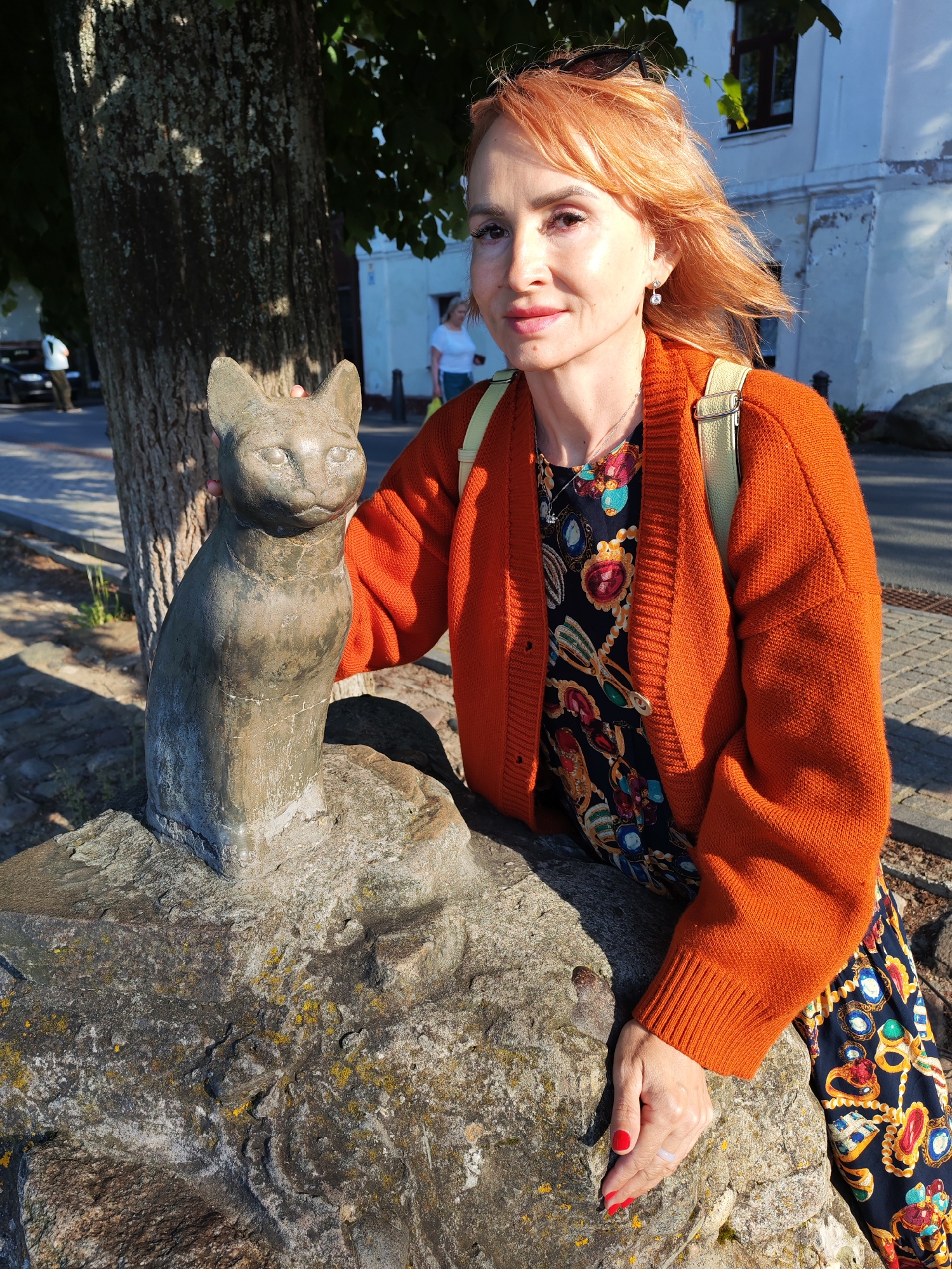 A woman with auburn hair and bright makeup stands in front of a stone cat sculpture on a rocky base. She is wearing an orange knitted cardigan over a dark dress with a colorful jewel-like pattern. A light-colored backpack is visible over her left shoulder. Her right hand rests on the sculpture. In the background, a tree trunk is prominent, and beyond that, a white building with a dark window can be seen. A paved walkway and some distant figures suggest an outdoor public space. The scene appears to be in the city of Plyos, Russia, possibly near the Museum-Reserve "Plyos" where a similar sculpture of a cat is located.