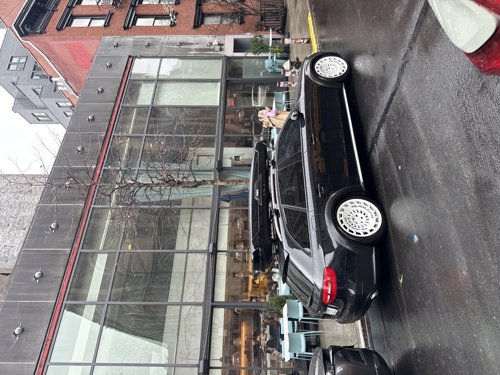 The image shows a street scene in New York City, United States of America. A black car with white wheels is parked on the wet asphalt street, next to a modern building with large glass windows. The building appears to be a cafe or restaurant, with people visible inside. A leafless tree stands in front of the building. Other buildings, constructed of brick and gray siding, are visible behind the modern building. Another car is partially visible behind the black car. The sky appears overcast.