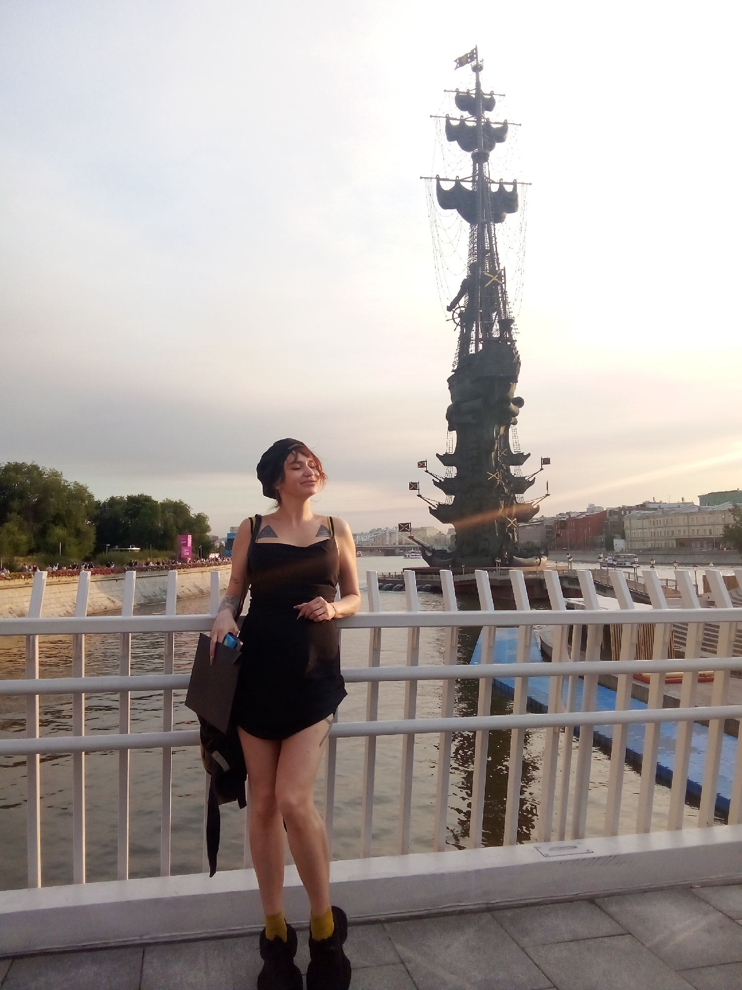 In an outdoor, urban setting, a woman stands on a paved walkway in front of a white railing. She is wearing a black dress, a black beret, and black furry slippers with yellow socks. She is holding a dark folder and a dark bag over her left shoulder. Behind her, a large statue of a ship with multiple masts stands in the water, and buildings can be seen in the distance. Trees line the opposite bank of the river. The sky is pale and overcast.