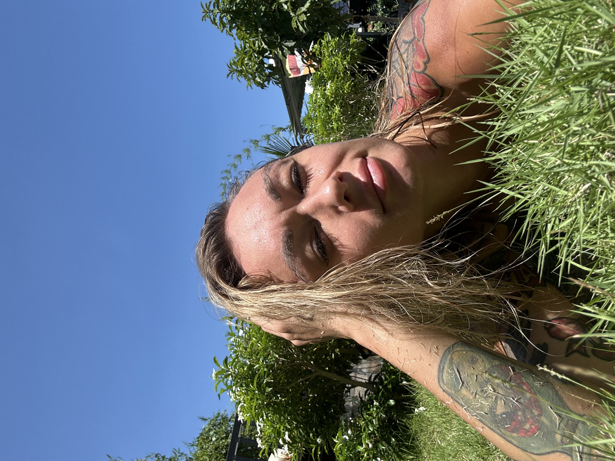 A woman is lying on her side in the grass under a clear blue sky. She is fair-skinned with blonde hair and has tattoos on her arms and shoulder. She has her eyes closed and her mouth slightly open. The background includes trees and a glimpse of a building. This photograph might have been taken in Mũi Né, Vietnam, considering the location request.