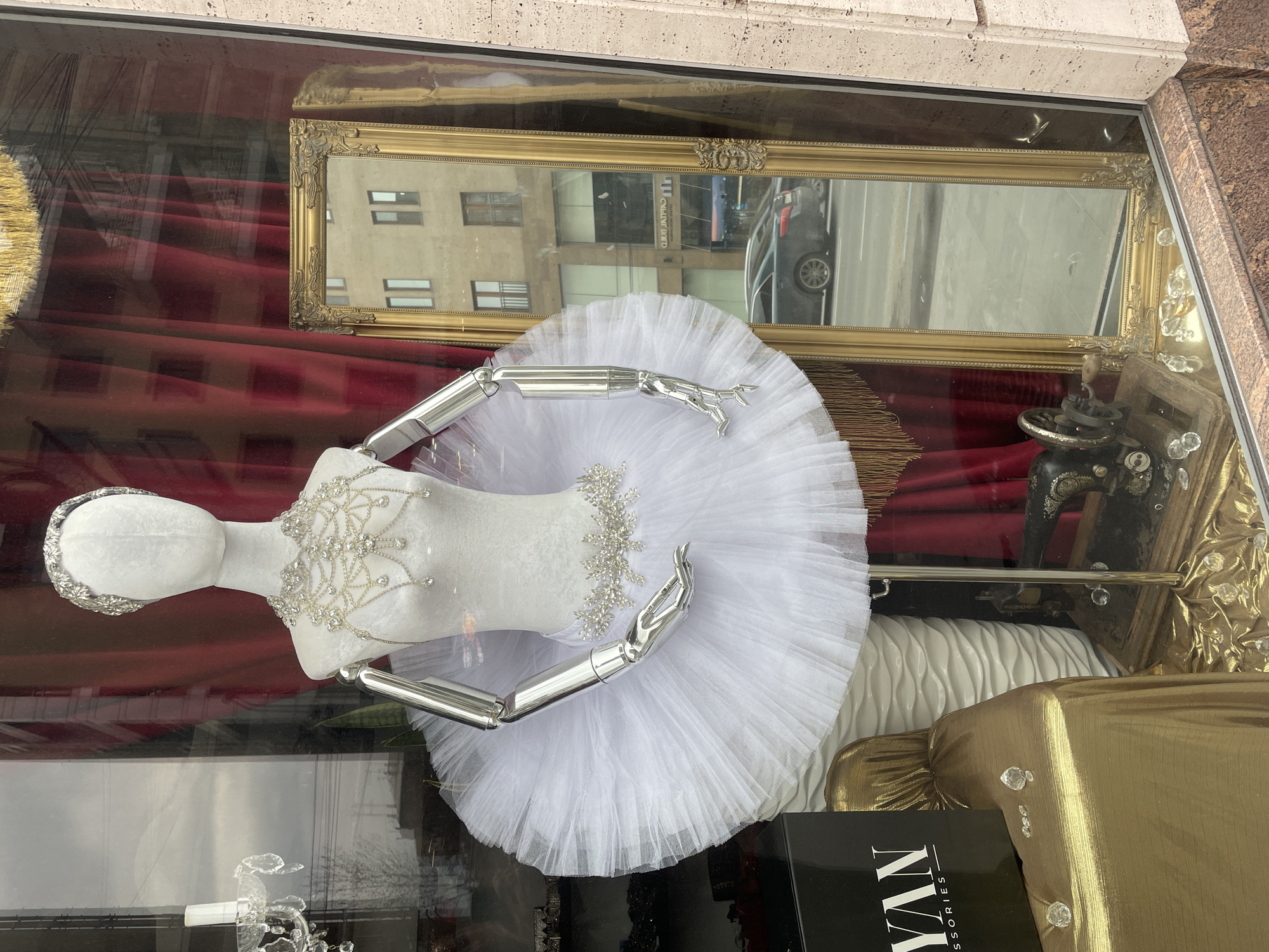 A storefront display in Yerevan, Armenia, showcases a ballerina mannequin in a white tutu and rhinestone-studded bodice. The mannequin has metallic arms and headwear, situated in front of a red curtain, a gold-framed mirror reflecting a street scene, and an antique sewing machine. A gold fabric draped over a box is visible near the base of the display, with "YAN Accessories" partially visible on the box.