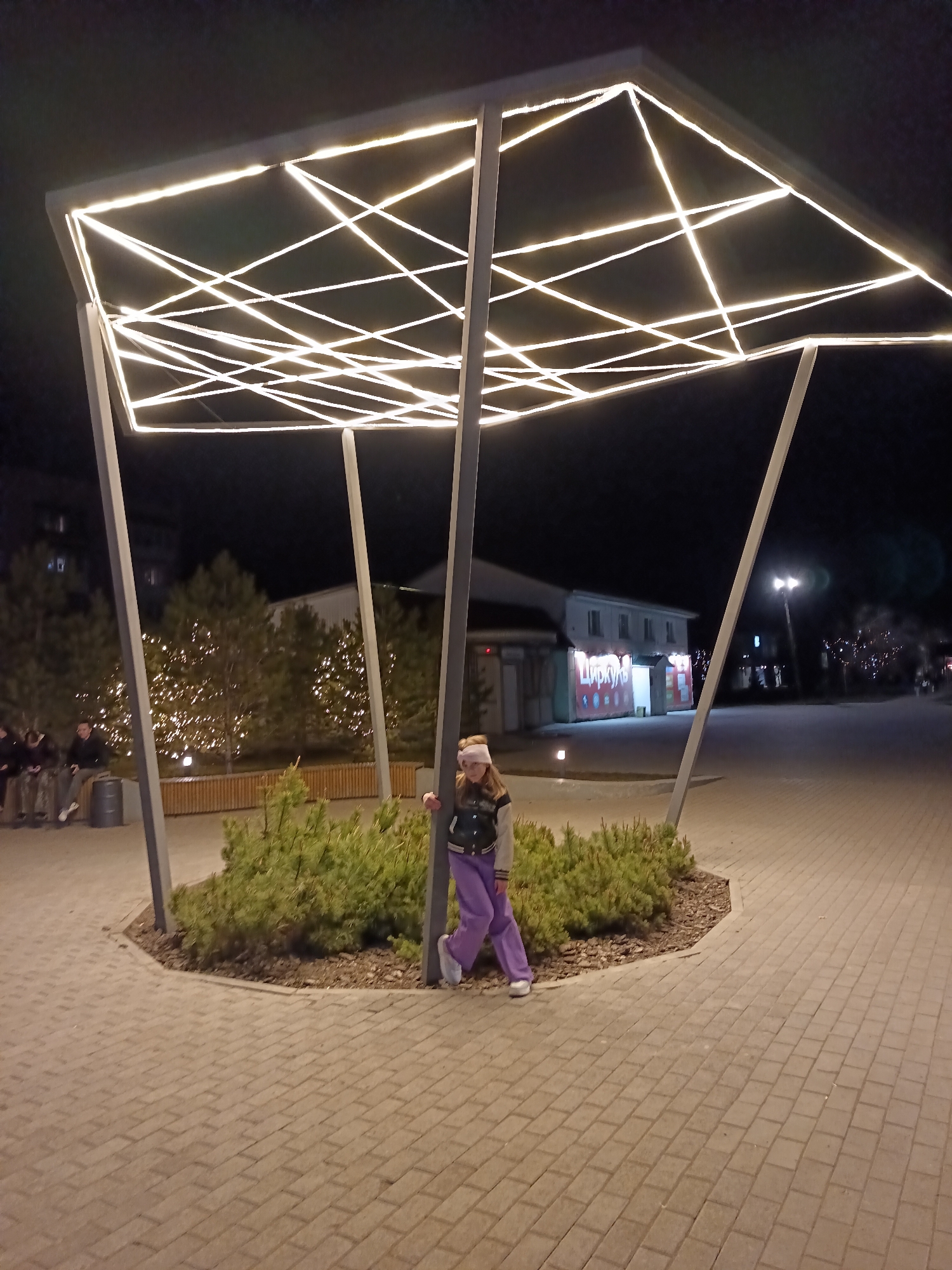 A full shot captures a girl in purple pants and a jacket leaning against a support of a decorative, illuminated structure in a public space in Osa, Russia. The structure has a flat, rectangular top outlined with bright white lights, with more lights creating a geometric pattern underneath. The structure is supported by metal beams. The girl is fair-skinned and wearing a headband. Behind her, a small, circular garden area is planted with shrubs. In the background, other people are seated on a bench under trees decorated with lights. Across a paved area, a building with Russian signage is visible. The ground is paved with rectangular paving stones. The scene is illuminated by artificial lights and appears to be nighttime.