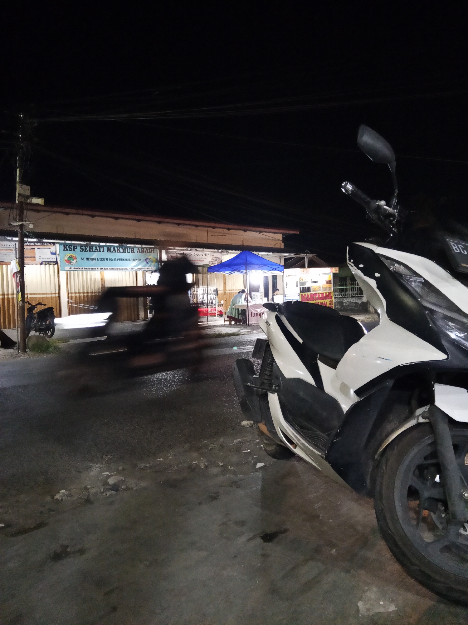 A night scene captured in Lubuklinggau, Indonesia, features a white and black motorcycle parked in the foreground, positioned to the right of the frame. Its front wheel and part of the body are visible. Behind it, a blurred motion of another motorcycle or vehicle is captured, suggesting movement. The background reveals a row of businesses or stalls. One prominent stall has a blue awning and displays food items. Another stall has a sign that reads "MARTABAK." The area appears to be an urban street or market at night, illuminated by artificial lighting. Power lines are visible overhead.