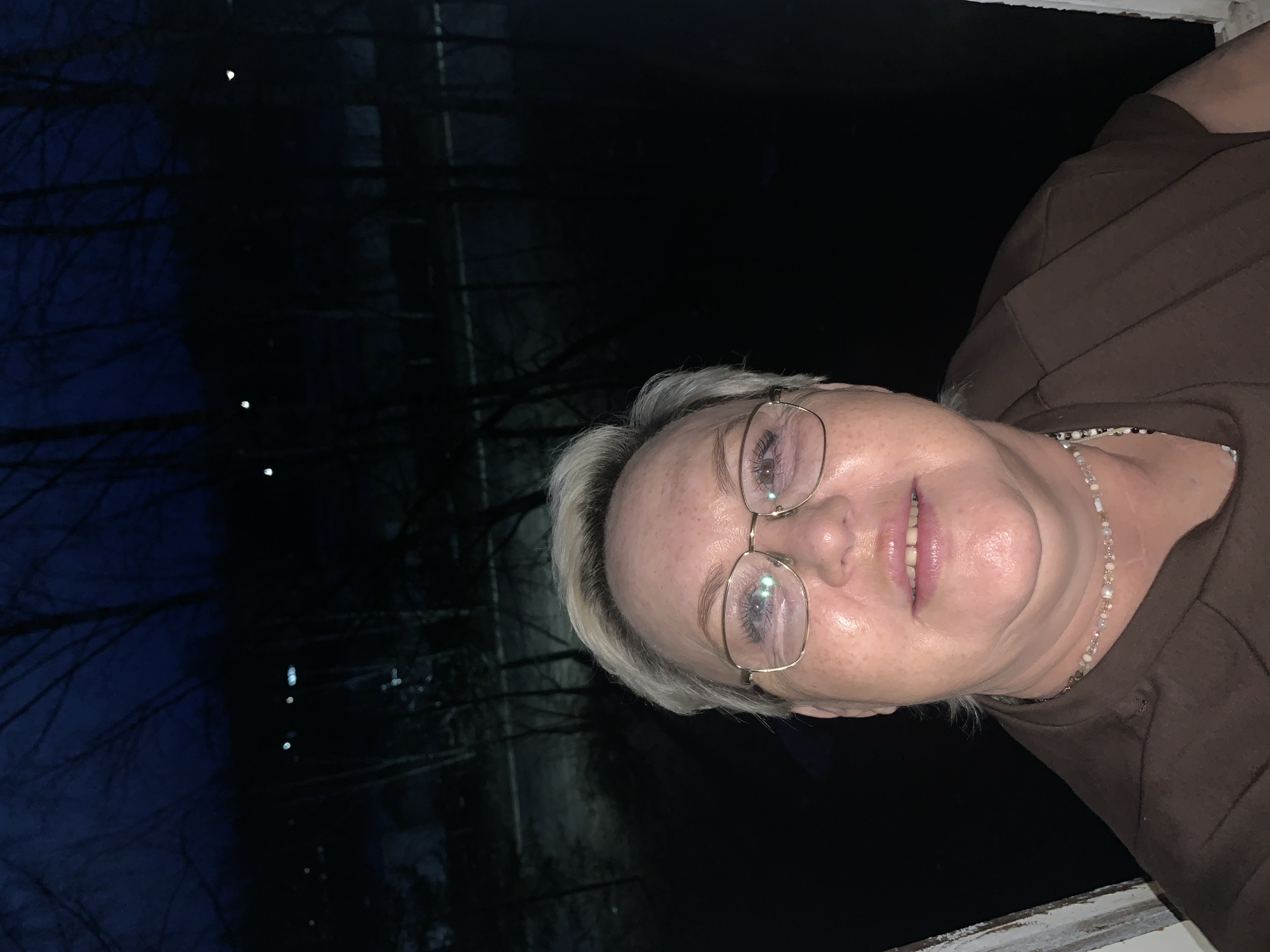 A woman with short, gray hair and eyeglasses stands in front of a dark window. Outside, dark trees are visible with sparse illumination. It's difficult to determine the location in Chaikovsky, Russia based on the image. The woman is wearing a brown shirt and a beaded necklace. She has a slight smile.
