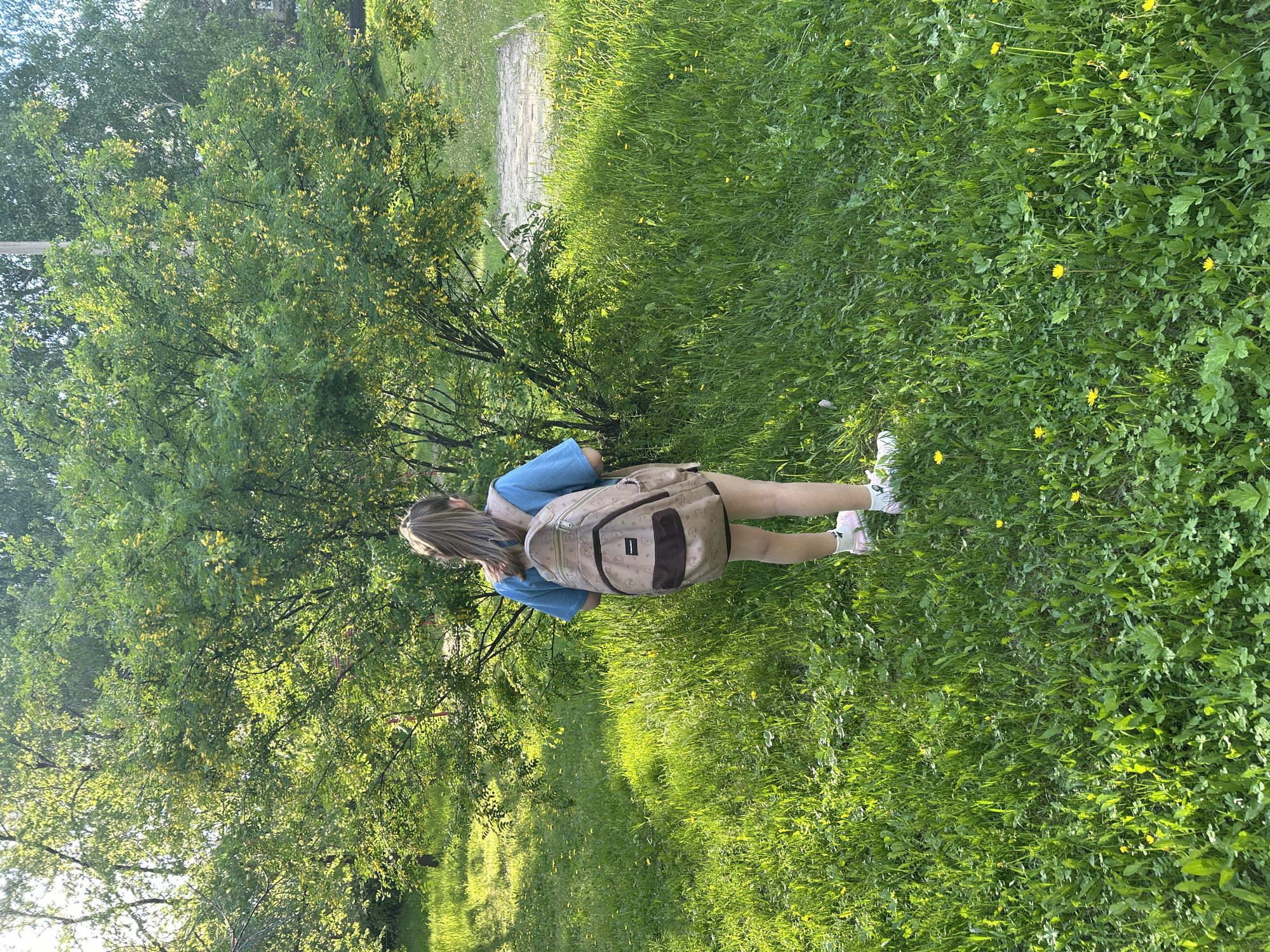 In this outdoor, eye-level shot, a young person with blonde hair is standing in a grassy area beside a tree. They are wearing a blue t-shirt, light-colored shorts, and white sneakers with pink accents. A light brown backpack with darker brown accents is slung over their shoulders. The backpack features a repeating pattern of cartoon animals.
The scene is a sun-drenched park or natural area. The sun is casting long shadows, indicating it is either early morning or late afternoon. The grass is vibrant green and dotted with small yellow flowers, likely dandelions. The tree to the left is full of lush green leaves, with some small yellow blossoms also visible. Beyond the grass, a stone pathway or paved area can be seen.
The overall mood is serene and peaceful, with the bright sunlight and natural setting contributing to a pleasant atmosphere. The person appears to be enjoying a moment outdoors. No visible text is present in the image. The location is identified as Angarsk, Russia.