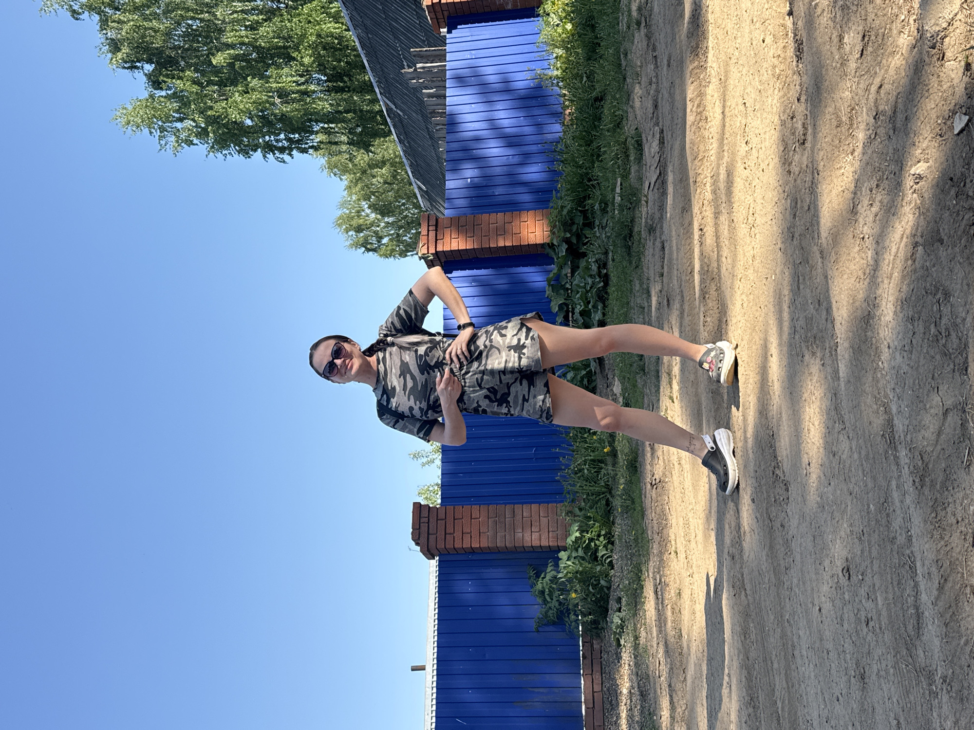 A young woman stands outdoors on a dirt path near a blue corrugated metal fence punctuated by brick pillars. She wears sunglasses and a camouflage print dress, with athletic shoes. The location is Rusalevka, Russia. A tree stands to the left of the fence, and the sky is clear and blue.