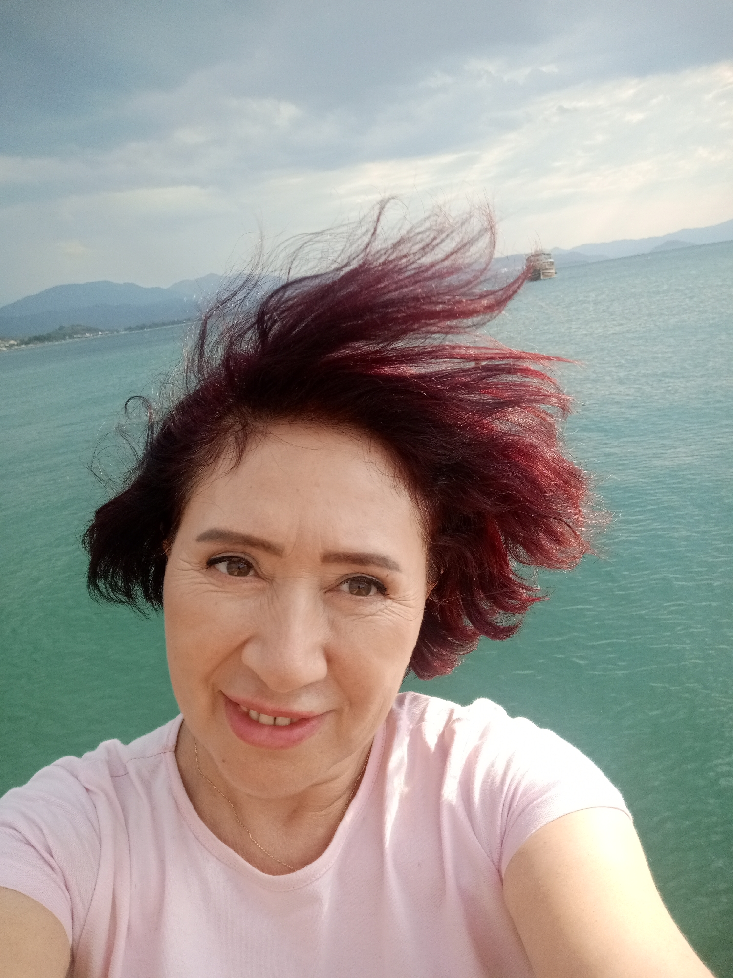A selfie is taken from a low angle outdoors. The subject is a middle-aged woman with short, dark red hair that is windswept. She has light skin, brown eyes, and is smiling with her teeth showing. She is wearing a pink t-shirt and a thin gold chain. The background is a large body of teal-colored water, with a boat visible in the distance. Mountains can be seen on the horizon. The sky is overcast with some patches of blue. The image appears to have been taken from a pier or waterfront location in Fethiye, Turkey.