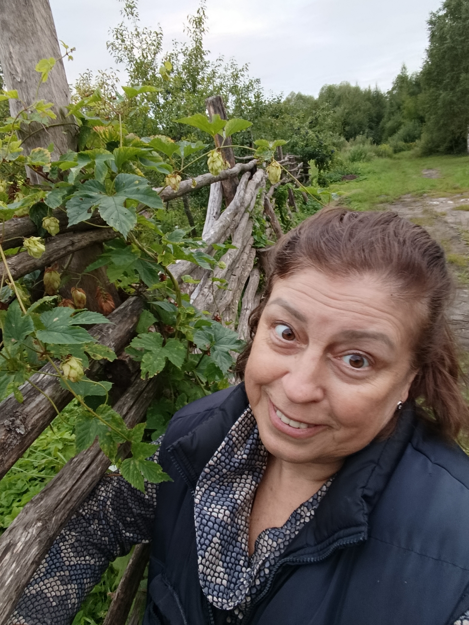 A selfie taken outdoors, with a woman in the foreground. She has brown hair and is looking at the camera with wide eyes and a smile. She is wearing a dark blue puffer jacket over a patterned scarf. Behind her, a rustic wooden fence is covered in green foliage and what appear to be hop cones. The background shows a dirt path and a dense line of trees under an overcast sky. The image captures a natural, rural setting, likely in the city of Vereya, Russia, given the context of the provided information.