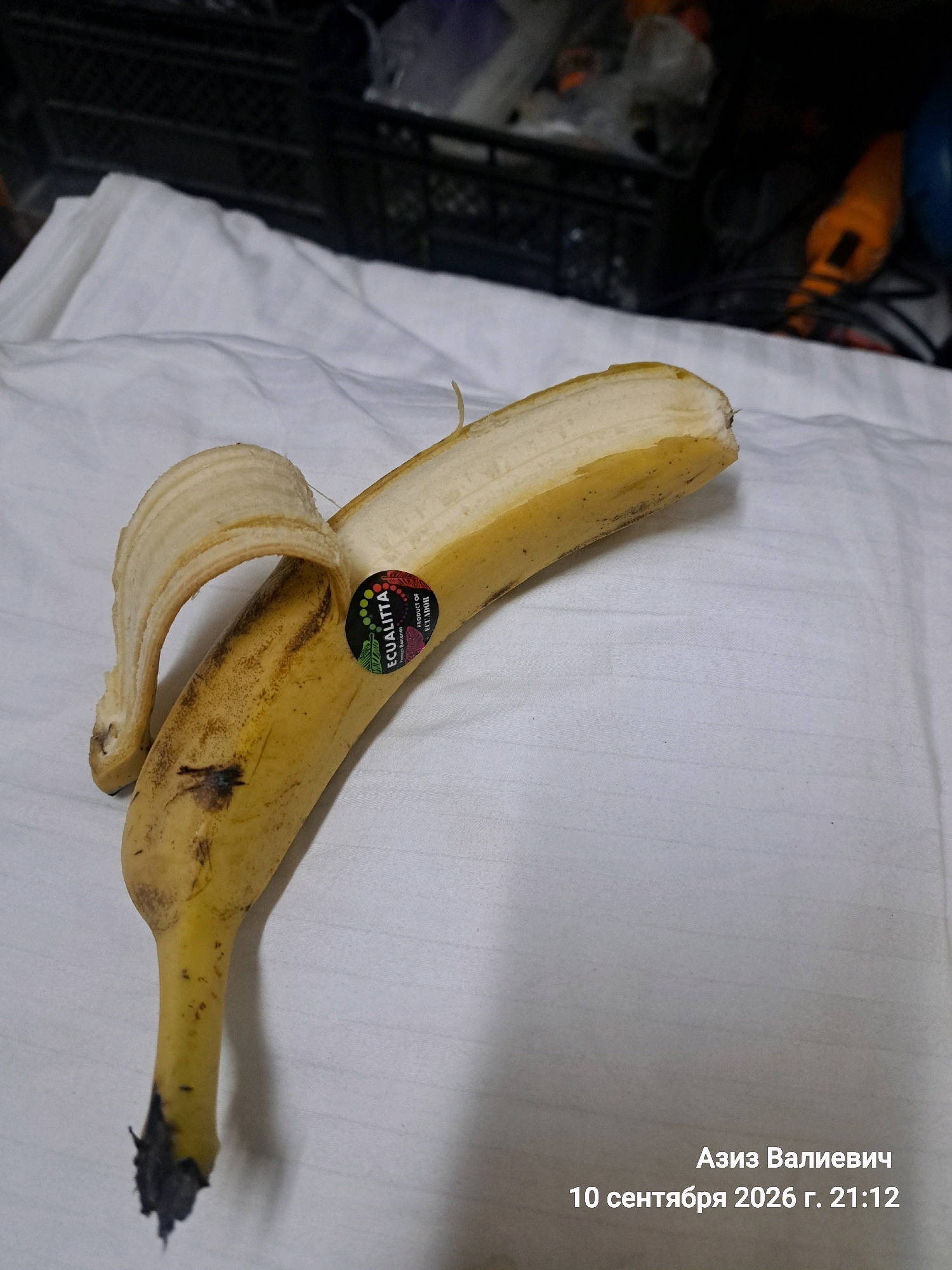 The image shows a partially peeled banana on a white, textured surface. The banana is yellow with brown spots, indicating ripeness. A sticker with the word "EQUALITTA" is visible on the skin. In the background, there appears to be a black grate and some blurred objects, suggesting an indoor setting. The bottom right corner of the image contains text in Russian, which translates to "Aziz Valievich" and "September 10, 2026, 21:12". There is no indication of the city of Tashkent, Uzbekistan in the image itself.