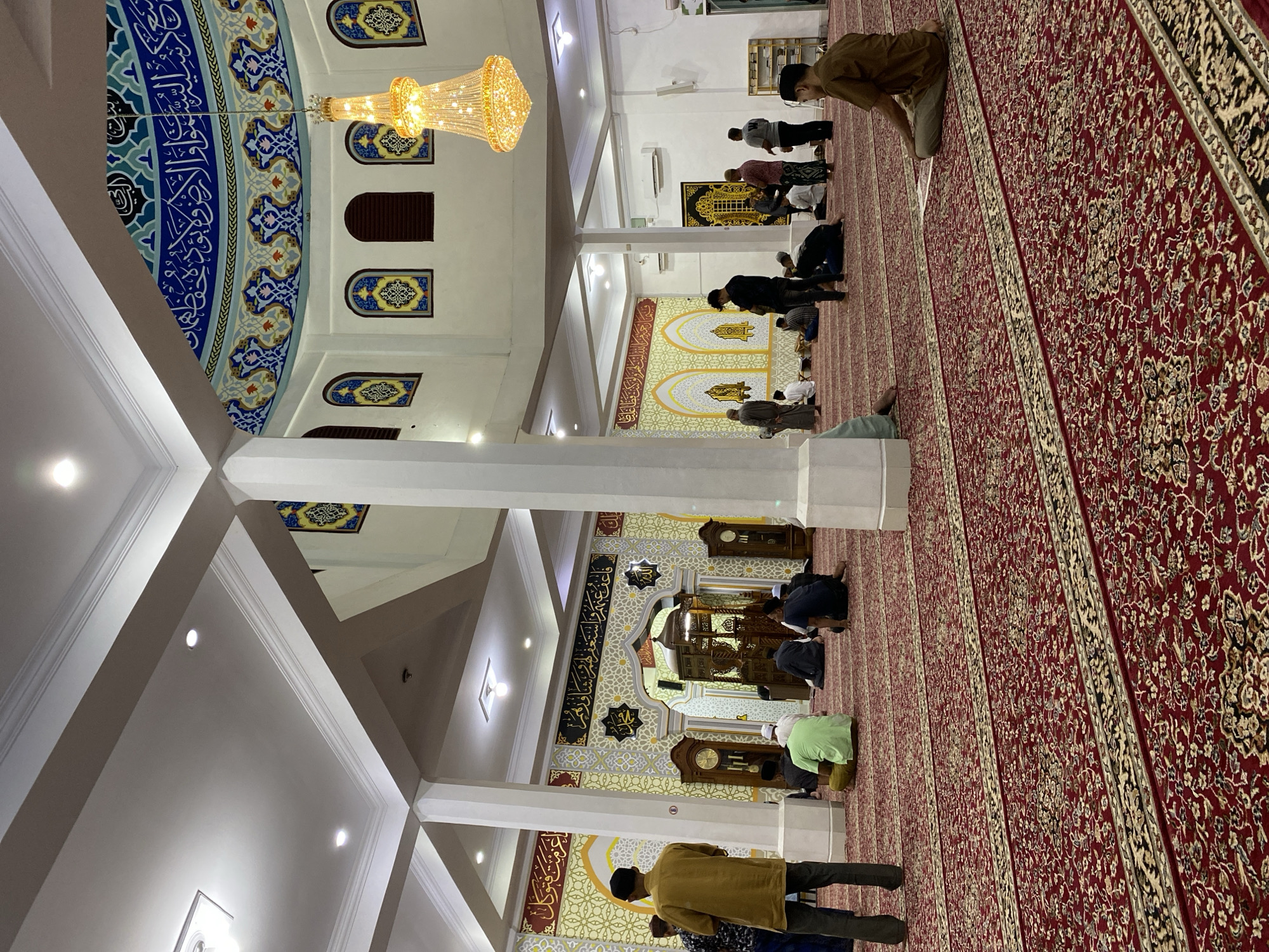 The image depicts the interior of a mosque, likely in Lhokseumawe, Indonesia, during prayer time. The scene is dominated by a large, ornate red carpet covering the entire floor. Several men are seen engaged in prayer, either standing or kneeling on the carpet. The mosque features high ceilings with intricate architectural details, including columns and decorative Islamic calligraphy on the walls and dome. A large, elaborate chandelier hangs from the ceiling, illuminating the space. The overall atmosphere suggests a moment of communal worship and tranquility.