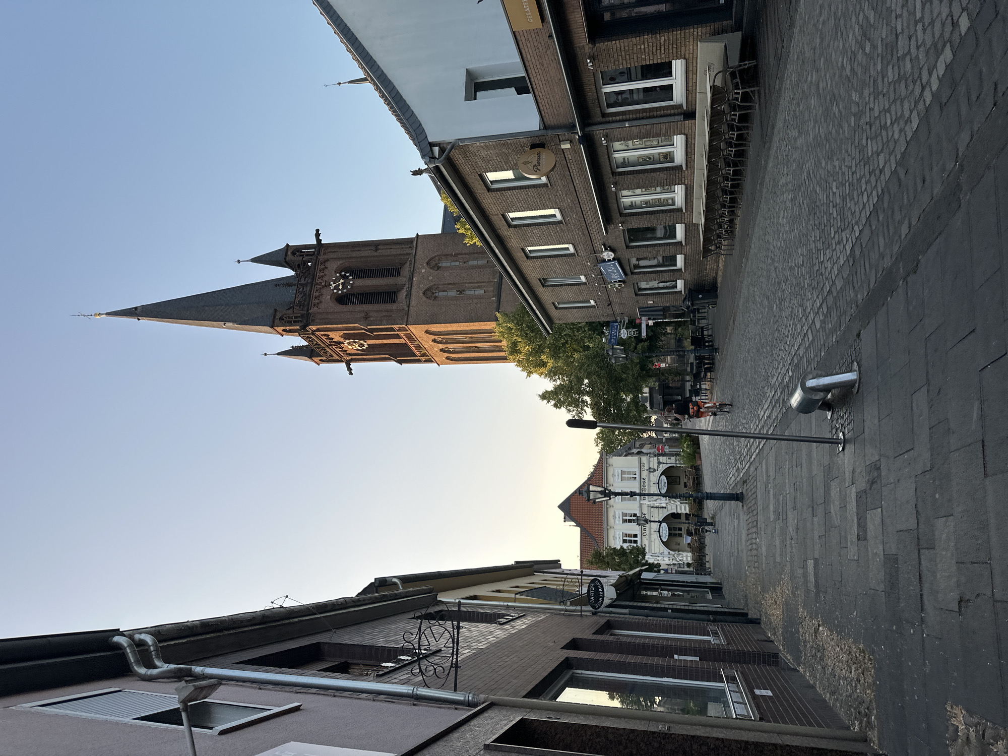 The image depicts a street scene in Krefeld, Germany, dominated by a large brick church tower with a pointed spire. The tower has a clock face. The street is paved with cobblestones and has sidewalks on both sides. Buildings line the street, some appearing to be shops with large windows. Outdoor seating with tables and chairs is visible in front of some establishments. A person is riding a bicycle down the street. The sky is clear and blue. The overall atmosphere suggests a late afternoon or early evening setting due to the warm lighting.
