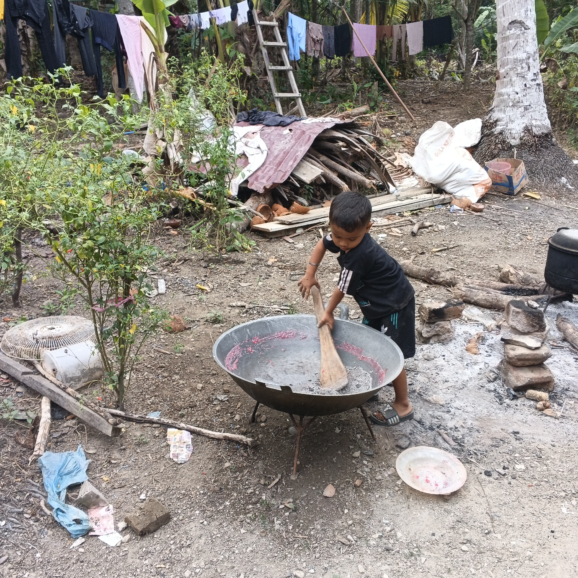 An exterior, medium shot captures a young boy, positioned in the lower right quadrant, standing over a large, metal wok. He is wearing a black t-shirt with white stripes on the sleeves and black shorts. His hands are engaged in stirring the contents of the wok with a wooden utensil. The wok rests on a three-legged stand, and ash and remnants of a fire are visible on the ground beneath it.
The surrounding environment appears to be an outdoor, somewhat unkempt area. In the background, laundry hangs on a line suspended between trees, with a wooden ladder leaning against a stack of firewood and debris. A large, rough-barked tree trunk is visible on the right, and vegetation, including a small chili plant with red peppers, is present in the foreground on the left. Several objects are scattered on the ground, including a white bag with text that appears to be "GULA KRISTAL", a discarded fan cage, a blue plastic bag, a small red bowl, and pieces of wood and coconut shells. The ground is dirt and gravel.