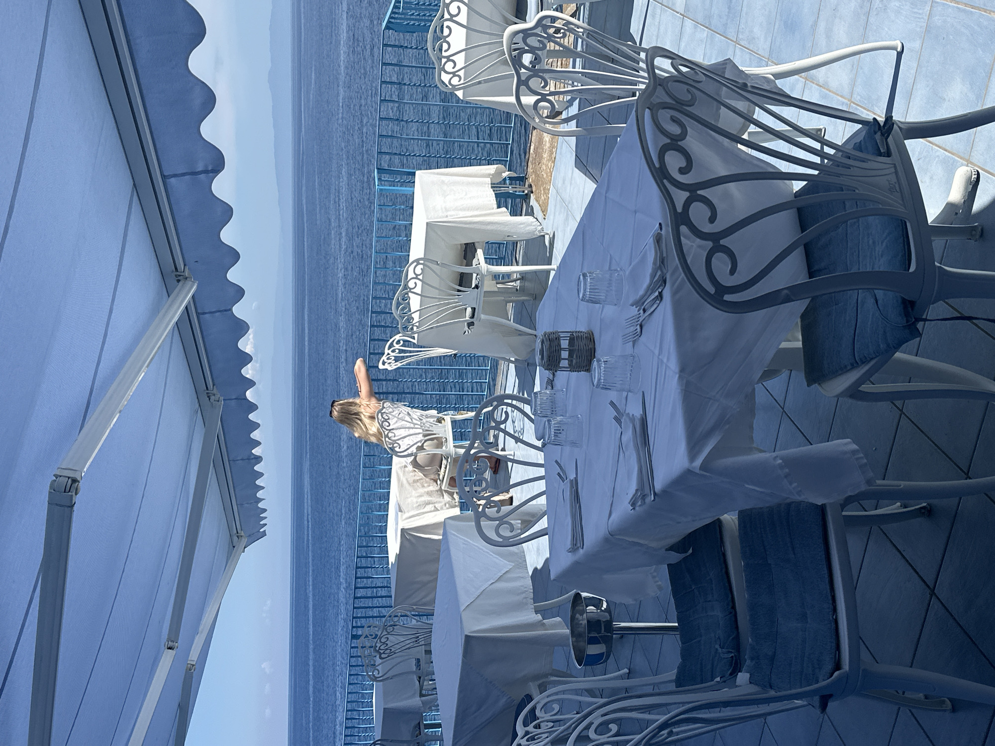This image captures a serene outdoor dining terrace overlooking the sea, likely in Lipari, Italy, given the context. The scene is dominated by shades of blue and white.
In the foreground, an expansive blue awning with a scalloped edge extends across the top, casting shadows on parts of the terrace. Several white tables are neatly arranged, each covered with a pristine white tablecloth. Around these tables are elegant white wrought-iron chairs featuring decorative scrollwork, some with blue fabric cushions. On the table closest to the viewer, a place setting is visible, including clear glasses, silverware, and a small, possibly woven, decorative object. A silver ice bucket on a stand is positioned near one of the tables. The terrace floor is tiled in a light blue.
In the mid-ground, a person with long blonde hair, seen from the back, is seated at one of the tables, facing the sea. They appear to be relaxing or enjoying the view. A blue metal railing runs along the edge of the terrace, separating it from the water below.
The background features the vast, calm blue sea, stretching to meet a clear, light blue sky on the horizon. Distant land or islands can be seen faintly in the far background. The bright natural light and shadows indicate a sunny daytime setting, suggesting pleasant weather. The overall atmosphere is one of tranquility and leisure at a picturesque coastal location. No visible text is present in the image.