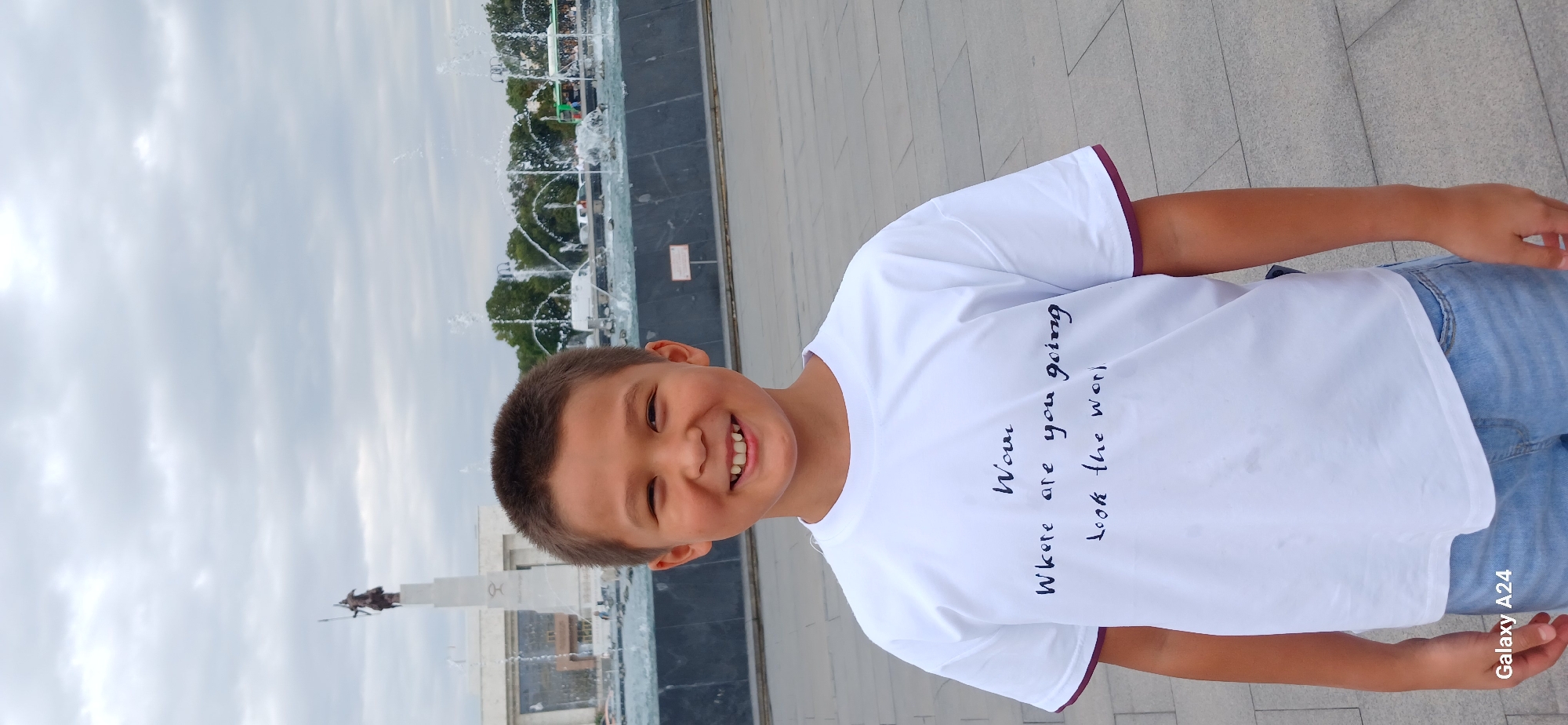 A young boy with short dark hair and a wide smile is standing in front of a fountain and a monument in Bishkek, Kyrgyzstan. He is wearing a white t-shirt with dark blue lettering that reads, "Wow, Where are you going, Look the world". He is also wearing light blue denim shorts. The monument in the background appears to be a statue of a person on horseback. The sky is overcast.