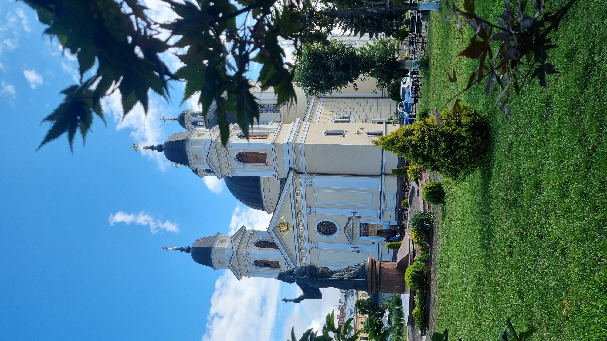 The image shows the Chernivtsi Holy Spirit Cathedral, a prominent landmark in Chernivtsi, Ukraine. The cathedral features a large central dome and two bell towers topped with crosses. In front of the cathedral is a statue of a robed figure holding a cross, placed on a pedestal. The foreground consists of a well-maintained lawn with manicured bushes and trees. In the background, cars are parked, and there are indications of other buildings and greenery, suggesting an urban setting. The sky is blue with scattered white clouds, and sunlight illuminates the scene, casting shadows. Several people are visible near the entrance of the cathedral and in the background, implying public access. The overall impression is one of a grand religious structure set within a pleasant, public space.