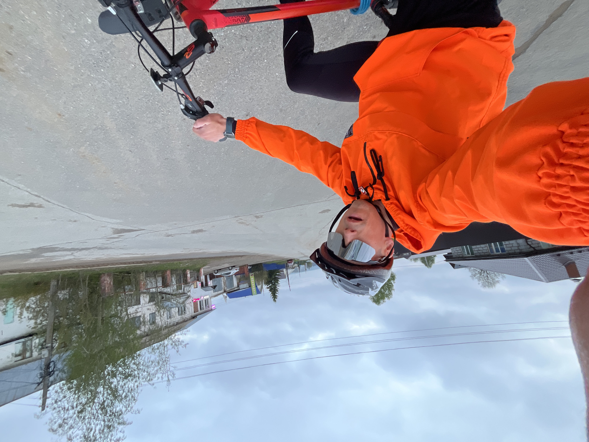 A person is riding a bicycle on a paved street in what appears to be the city of Chaikovsky, Russia. The person is wearing a bright orange jacket, black cycling pants, a helmet, and sunglasses. They are holding the handlebars of a red and black mountain bike. The background shows a cloudy sky and various buildings, including older, multi-story residential buildings on the left and more modern houses on the right. There are trees present along the street. The perspective suggests the photo was taken by the person riding the bike.