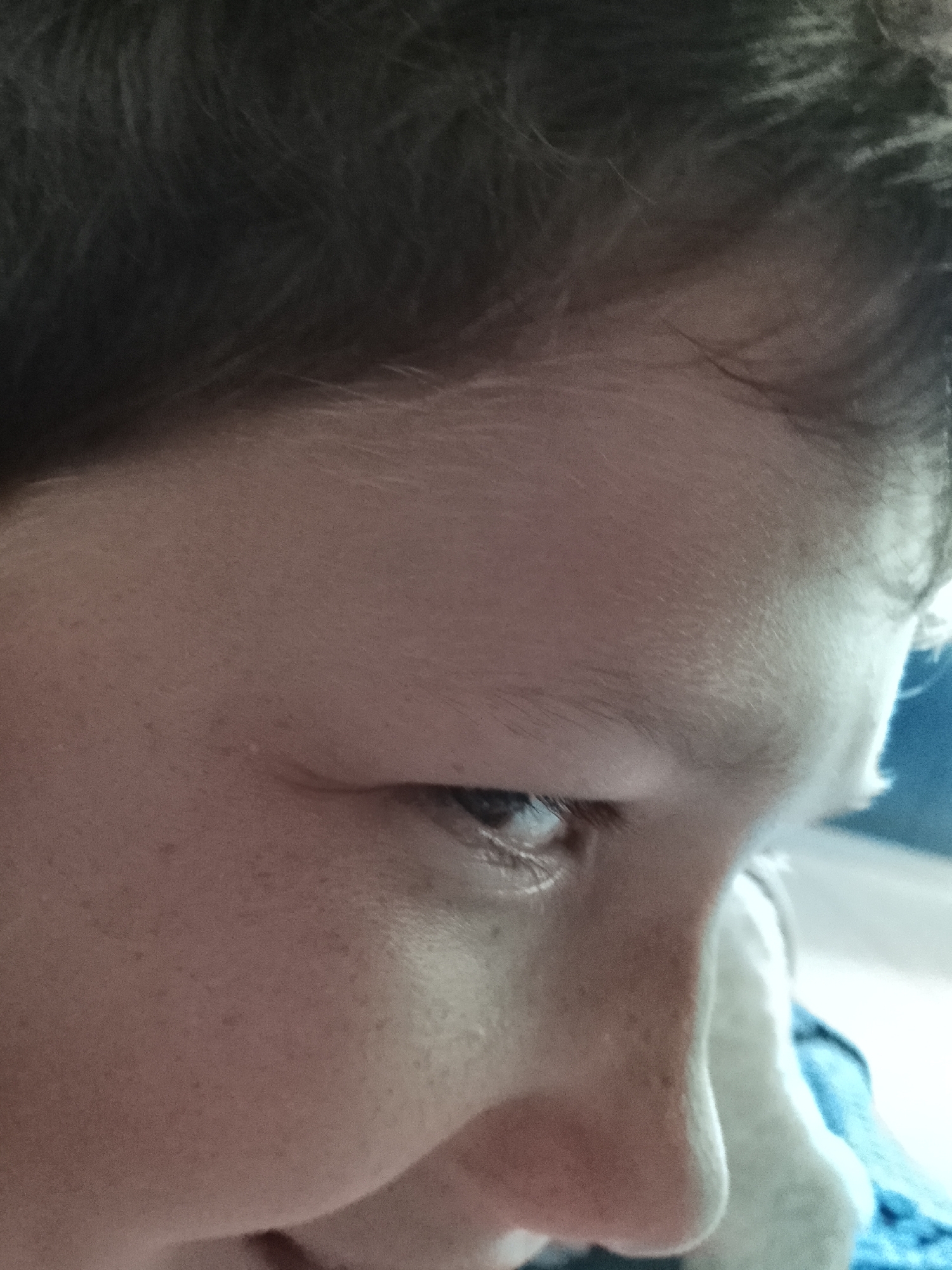 The image is a close-up, low-angle shot of a person's face, focused on the left eye and cheek. The individual's eye is visible, with eyelashes and the iris appearing dark. The skin is fair with visible pores and some freckles. The eyebrow is dark and well-defined. The person's hair, which appears dark brown and slightly messy, frames the upper part of the face. The background is out of focus, with a blur of colors that suggest an indoor environment, possibly a bedroom or living area. There is a hint of blue fabric to the right of the face. The overall lighting is soft, casting gentle shadows. The image was taken in the city of Minino, Russia.