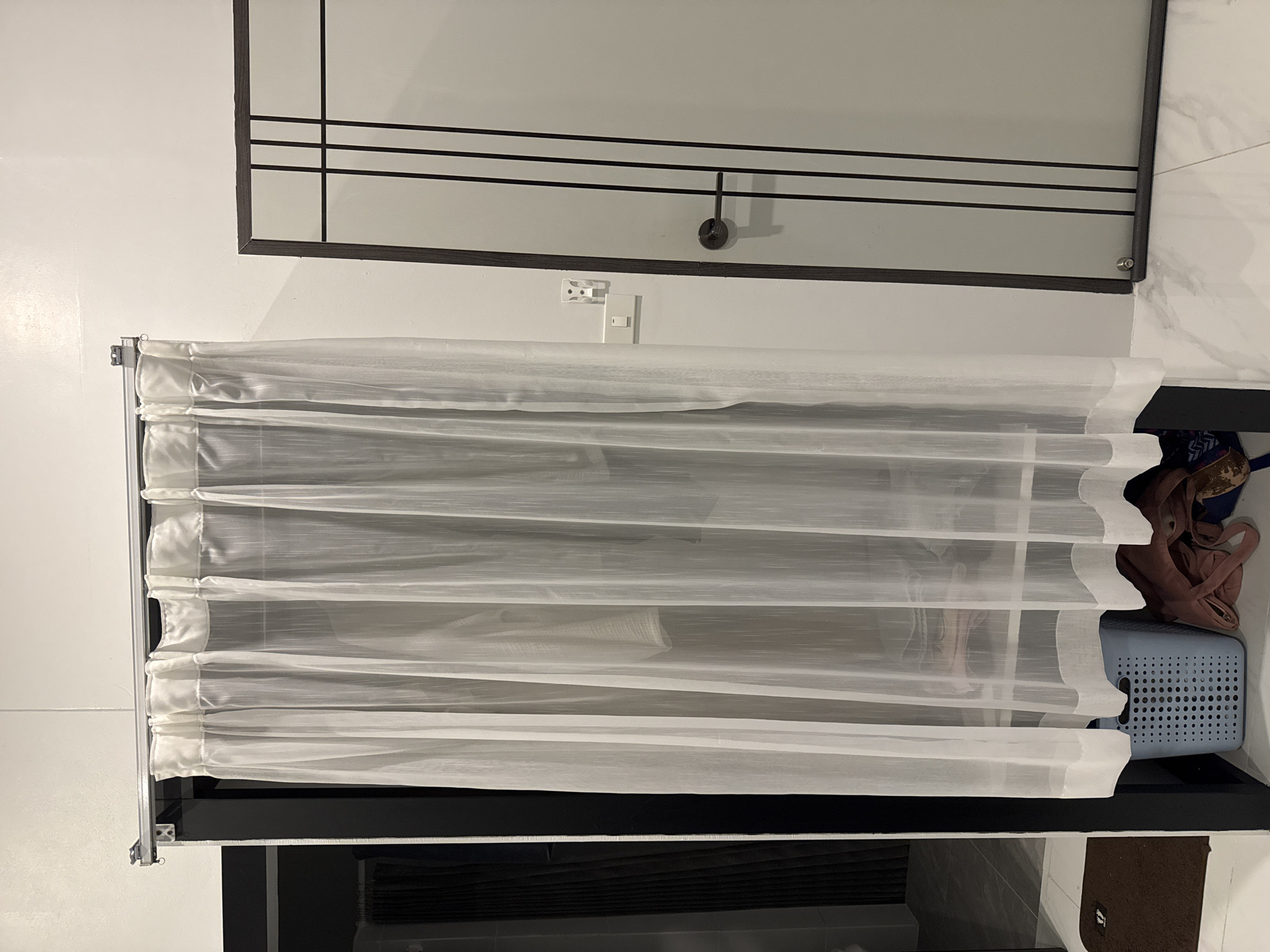 The image shows an interior scene, likely a closet or dressing area, in a room with white walls and light-colored tiled flooring. On the left, a white sheer curtain is hung from a metal rod, covering an open doorway or closet. Behind the curtain, some clothing and shelves are visible. To the right of the curtain is a light gray door with dark vertical stripes and a dark handle. A light switch and a white electrical outlet are mounted on the wall to the left of the door. On the floor in front of the curtain, there is a blue plastic laundry basket and some fabric items, including a pink bag. A dark brown bath mat is partially visible on the floor to the far left. The scene appears to be well-lit, suggesting indoor lighting. The location is specified as Tambon Koh Phangan, Thailand.