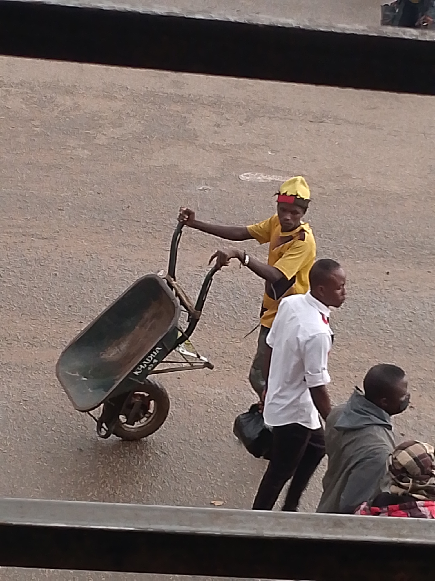 A medium shot captures a scene in Bukuru, Nigeria. A young man in a yellow t-shirt and a matching cap with red trim is pushing an empty wheelbarrow. The wheelbarrow has "KADUNA" printed on the side. A young man in a white shirt and black pants walks behind him. Another man in a gray jacket with a face mask and a person with a patterned scarf are also visible in the background. The setting is an outdoor area with an asphalt surface.