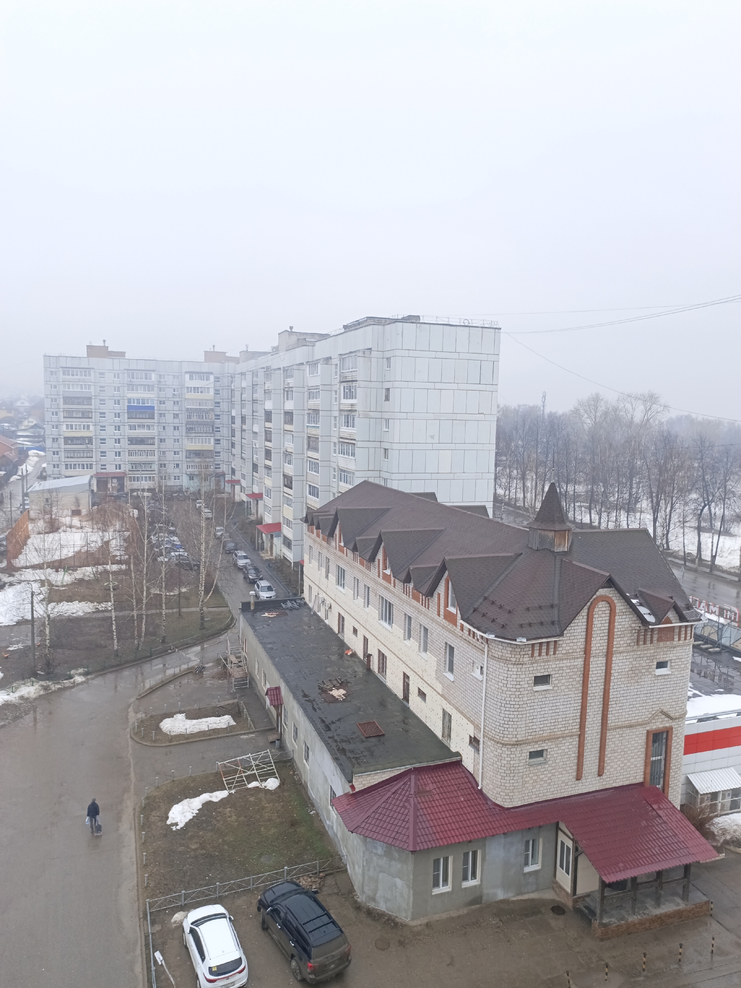 This image depicts an urban scene in Chaikovsky, Russia, characterized by a thick fog obscuring the background. Several multi-story residential buildings constructed of concrete slabs dominate the view, their facades a pale gray. A smaller, brick-faced structure with a dark brown gabled roof and a turret-like detail is positioned closer to the foreground. This building appears to have an adjacent low structure with a red tile roof.
Below the building complex, a wet asphalt street curves past, exhibiting signs of melting snow and slush. Bare trees line the street edge, contributing to the wintry ambiance. A single individual is walking along the street pulling what appears to be a suitcase on wheels. In the foreground, a couple of parked vehicles, a white sedan and a dark-colored SUV, are visible.
The scene conveys a sense of quietness and stillness due to the weather conditions and the lack of activity. The overall impression is that of a typical urban landscape in a cold, foggy climate.