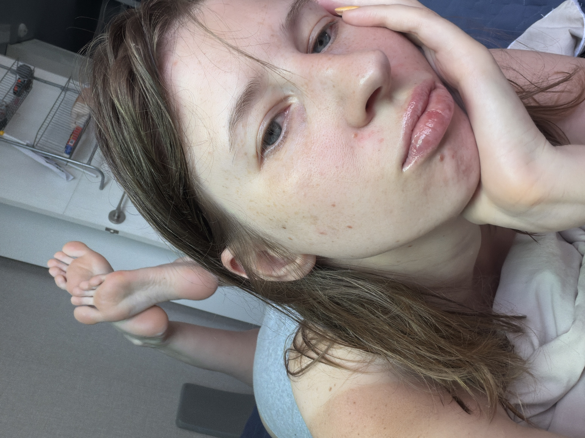 A young woman is lying on a bed in Dongducheon-si, South Korea. The scene is indoors, with a hint of daylight suggesting it is daytime. The woman is positioned in a way that her face, one foot, and part of her arm are visible. Her face is close to the camera, with her eyes open and her hand resting on her cheek. She has a light complexion with freckles and appears to be wearing lip gloss. Her hair is brown and slightly messy, framing her face. Her left foot is extended and bent at the ankle, with the sole of her foot facing the camera. Her toes are spread slightly. She is wearing a light-colored top and appears to be under a white blanket. In the background, there is a hint of a white dresser or cabinet and a metal rack with some items on it. The overall mood of the image is casual and relaxed. No visible text is present in the image.