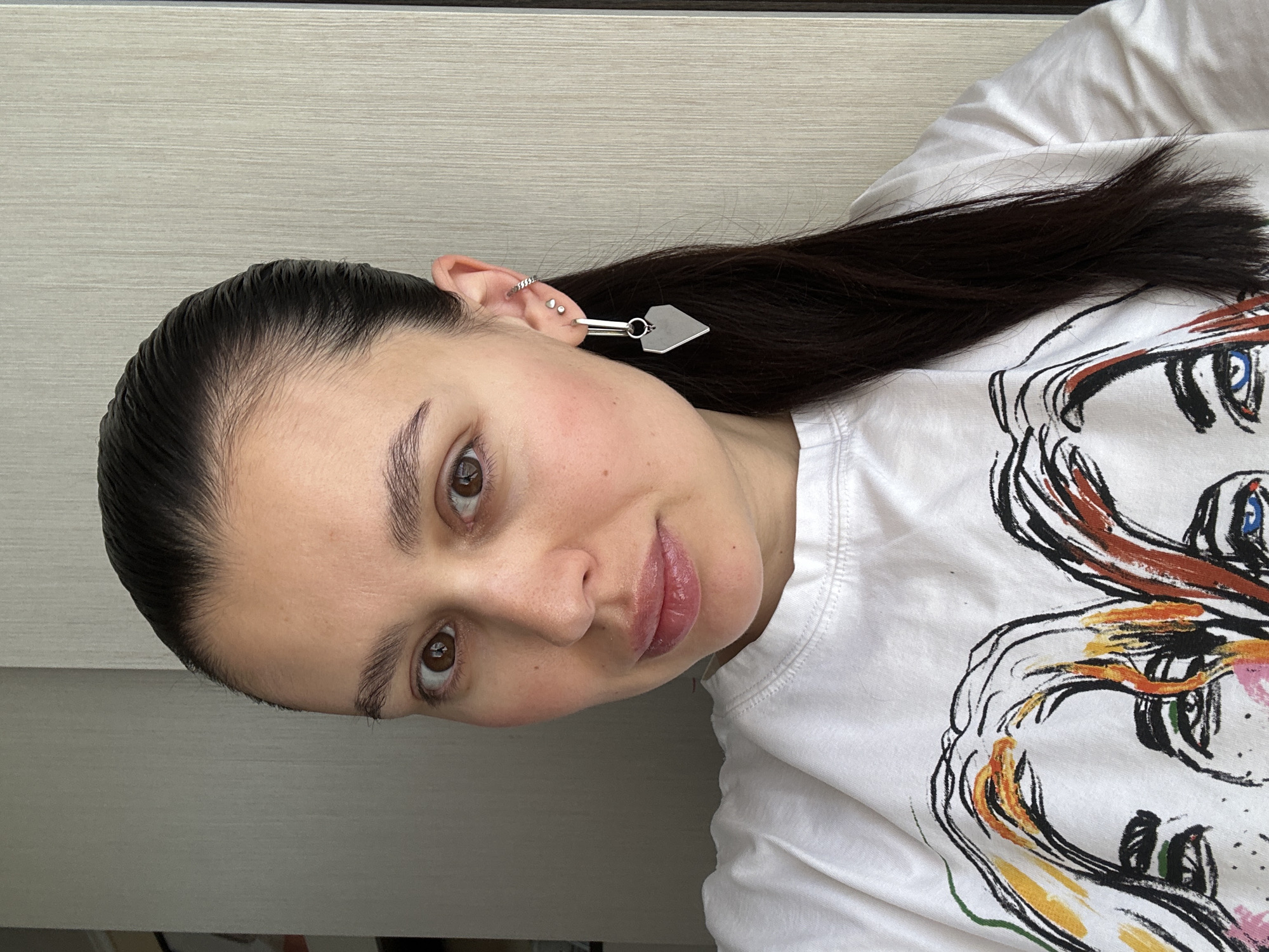 The image features a young woman in an indoor setting, likely in Chaikovsky, Russia, posing for a photograph. Her dark, sleek hair is pulled back tightly, showcasing her facial features and revealing fine baby hairs around her hairline. She looks directly at the camera with a calm, slightly inviting expression, characterized by her brown eyes, well-defined eyebrows, and subtly glossed lips hinting at a gentle smile.
A notable detail is her left ear, adorned with multiple silver piercings: a helix ring and two small studs. A distinctive silver earring, designed with a minimalist bar from which a flat, house or arrow-shaped pendant hangs, completes her accessory choice.
She is wearing a white crew-neck t-shirt that features a prominent, colorful, and abstract graphic print. The design resembles a bold sketch, possibly depicting an animal or a stylized face, utilizing strong black outlines filled with vibrant splashes of orange, blue, pink, and green. Her dark hair cascades over her shoulder, partially obscuring the graphic.
The background consists of a plain, light-colored wall with a subtle horizontal wood-grain or textured pattern. The lighting is soft and even, suggesting a daytime indoor environment without harsh shadows. The photograph is oriented at an angle, giving the impression that the subject might be reclining or that the camera was tilted during capture. There is no visible text in the scene.