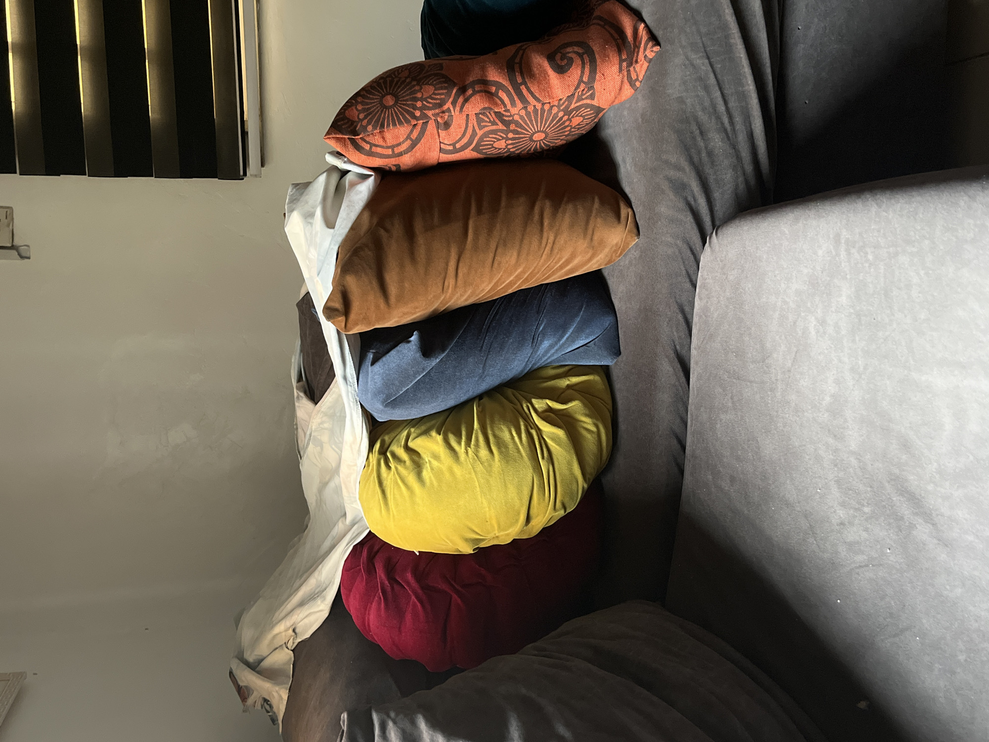 The image depicts a stack of six pillows of varying colors (orange, brown, blue, yellow, red, and white) placed on a grey couch. The location is possibly a home setting due to the presence of a window blind visible on the upper left corner and the general look of furniture. The city or country of Abuja, Nigeria cannot be confirmed from the visual information. The pillows appear to be resting on a cushion or similar surface, with the grey couch filling the frame. There are no individuals or interactive elements in the image.