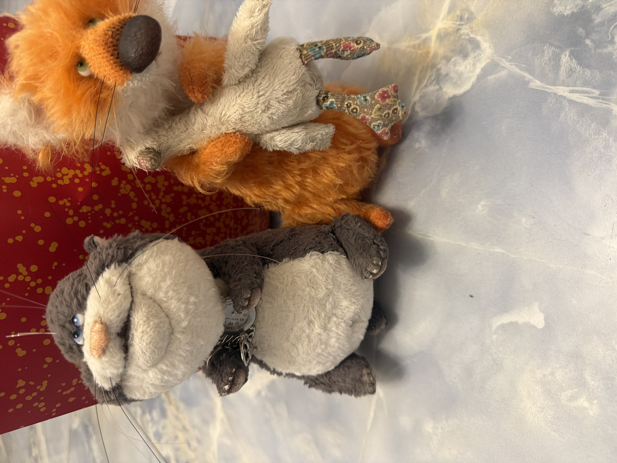 Two stuffed toys, a dark gray, round-faced animal with blue eyes and a prominent smile, and an orange, fluffy fox-like animal with green eyes, are positioned on a light blue, marble-patterned surface. The gray animal is on the left, wearing a silver chain with a round pendant around its neck. The fox is on the right, holding a light-colored stuffed bird in its arms and wearing patterned fabric on its legs. In the background, a red object with yellow speckles is visible. The lighting suggests an indoor environment.
