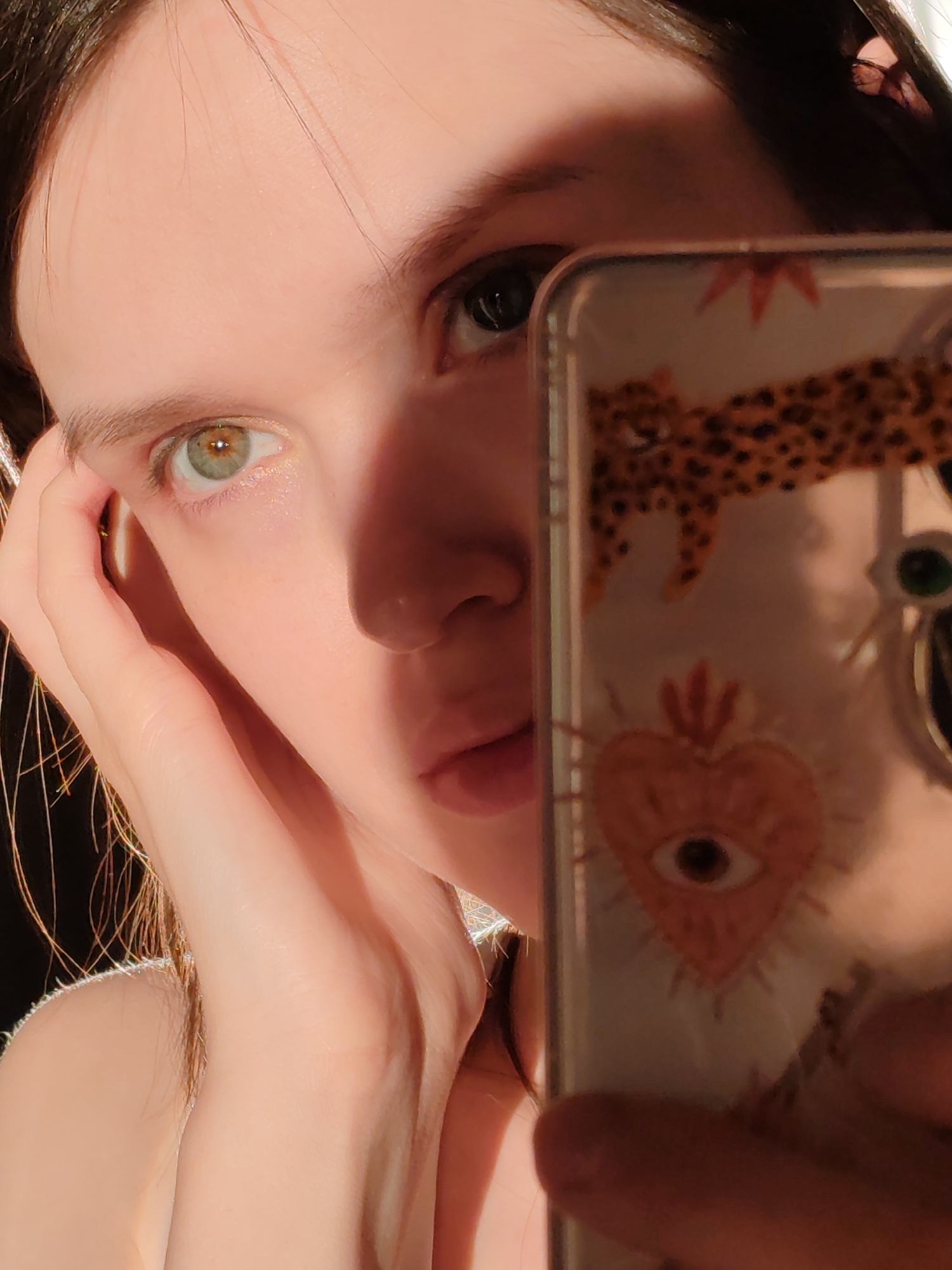 A fair-skinned person with heterochromia (different colored eyes) is holding a smartphone up to their face, partially obscuring it. The right eye is dark, while the left eye is lighter, possibly green or hazel. The person's skin is smooth, and their lips are slightly parted. Light falls across the left side of their face, creating a contrast with the shadowed right side. The smartphone case is decorated with a leopard print at the top and a heart with an eye inside at the bottom. The person's hand is near their face. The background is dark and out of focus. It is impossible to determine the location of Ust-Ilimsk, Russia, from this image.