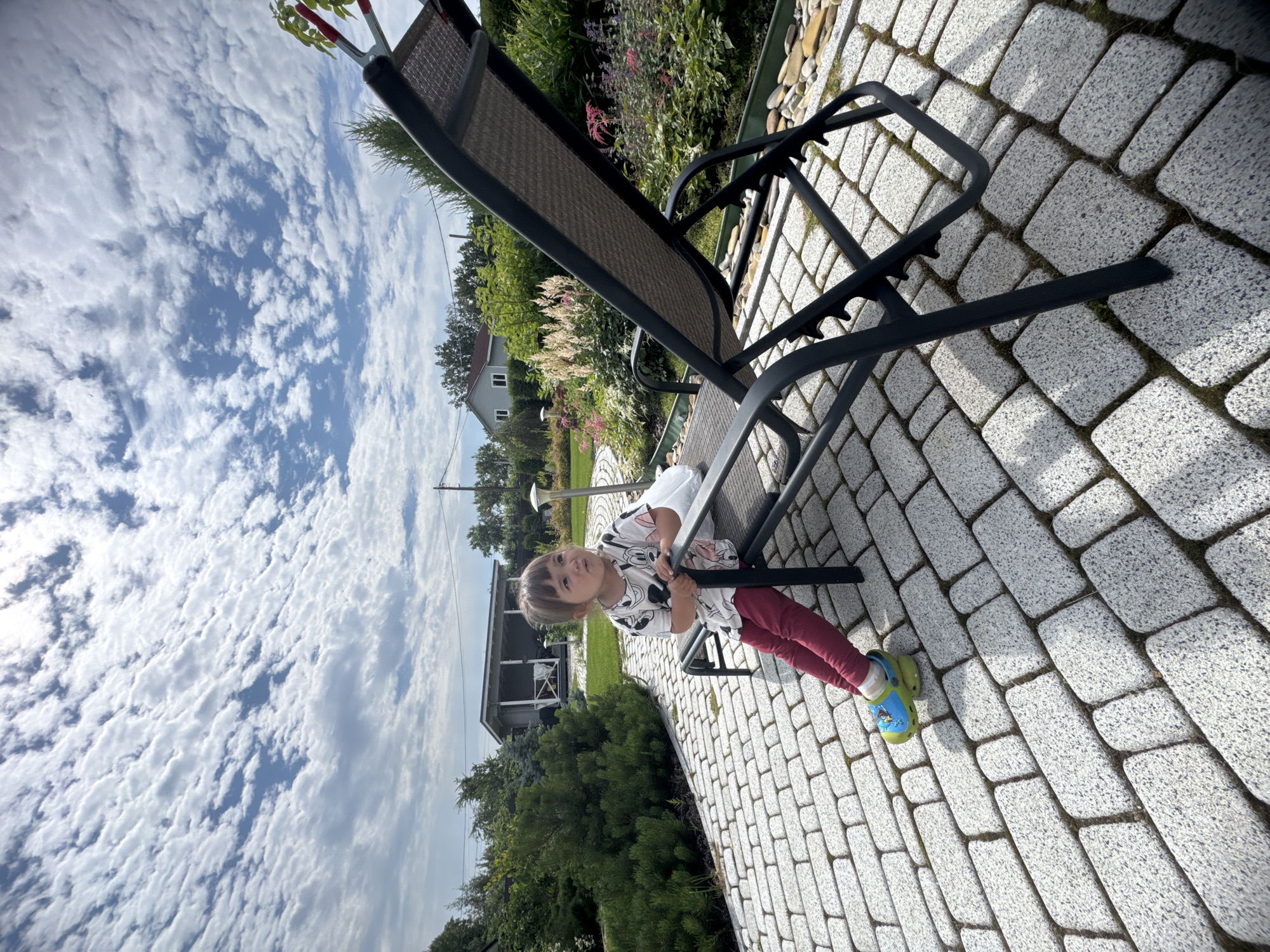 A low-angle, wide-shot photograph taken outdoors in the city of Oliki, Russia, shows a young child sitting on a dark gray reclining lawn chair. The child, who appears to be a toddler, is positioned on the left side of the chair, facing left and looking upwards. They are wearing a white t-shirt with a Mickey Mouse print, dark red leggings, and light green clogs with blue accents. The child's hands are gripping the armrest of the chair.
The lawn chair is situated on a patio paved with light gray rectangular cobblestones. The patio is bordered by a green lawn on the right, with various plants and flowers in bloom. In the background, several houses with white walls and dark roofs are visible, surrounded by lush green trees and bushes. The sky above is a vibrant blue, filled with dramatic, textured white clouds, suggesting a partly cloudy day. The sun appears to be casting shadows, indicating it is likely daytime. The overall scene depicts a peaceful, domestic outdoor setting.