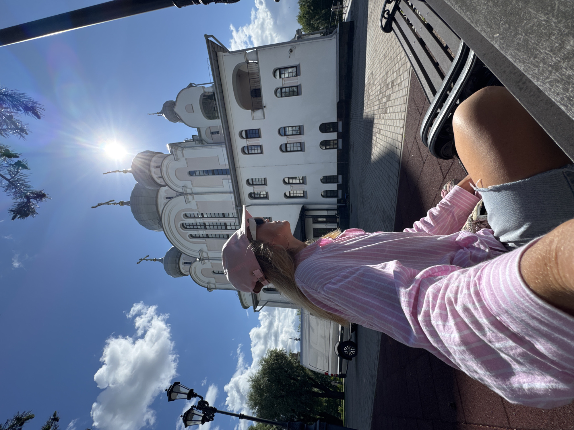 In Saint Petersburg, Russia, on a sunny day, a woman wearing a pink and white striped shirt, a pink baseball cap, and sunglasses sits on a park bench. Her legs are crossed, and she is looking to her left. Behind her, a white church with a pink trim and gray domes stands under a bright blue sky with scattered white clouds. The sun is shining brightly, creating lens flare in the image. In the background, there are trees, a streetlamp, and a silver van. The area in front of the church is paved with cobblestones and there is a red rubberized surface to the left of the bench.