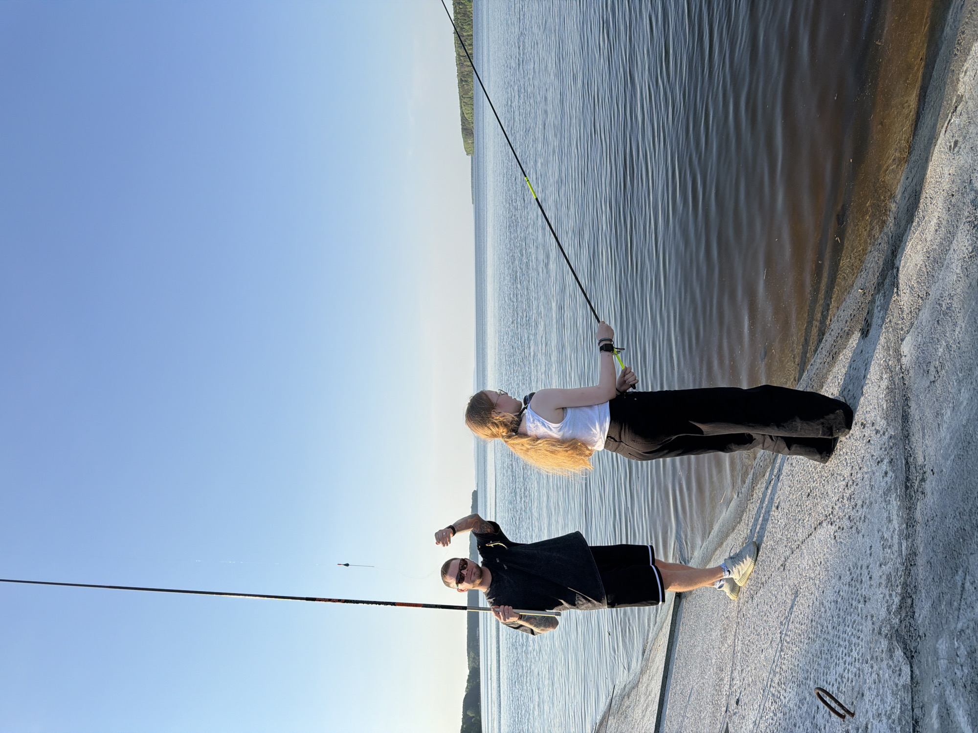 Two people are fishing near water under a blue sky. The image appears to be taken on a concrete structure, such as a pier or embankment. The person closer to the bottom of the image is a man with tattoos on his arms, wearing sunglasses, a dark-colored t-shirt, and black shorts with a white stripe. He is holding a fishing rod. The person near the top of the image is a woman with long blonde hair, wearing a white top and black pants. She is also holding a fishing rod. The water is calm with ripples. A shoreline with trees is visible in the distance. The photograph seems to be taken from a slightly low angle and rotated 90 degrees counterclockwise. It is difficult to ascertain specific geographic location without additional landmarks visible. Chaikovsky, Russia can not be determined.