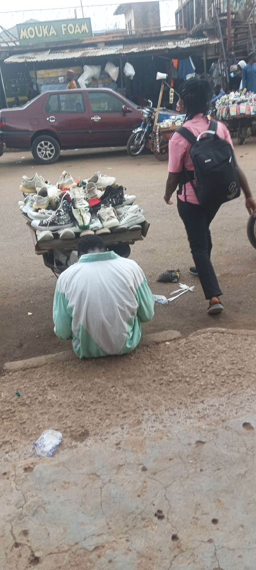 The image depicts a street scene in Bukuru, Nigeria. A man sits on the ground next to a wooden cart piled high with shoes for sale. The cart is positioned on a roadside next to a concrete barrier. A woman wearing a pink shirt, black pants, and a black backpack walks past the cart, carrying what appears to be a spare tire or wheel cover. In the background, a red car is parked in front of a shop with a sign that reads "MOUKA FOAM". A motorcycle is parked nearby. Other people and market stalls can be seen further in the background. Litter, including plastic wrappers and debris, is scattered on the ground. The overall scene suggests a busy commercial area.