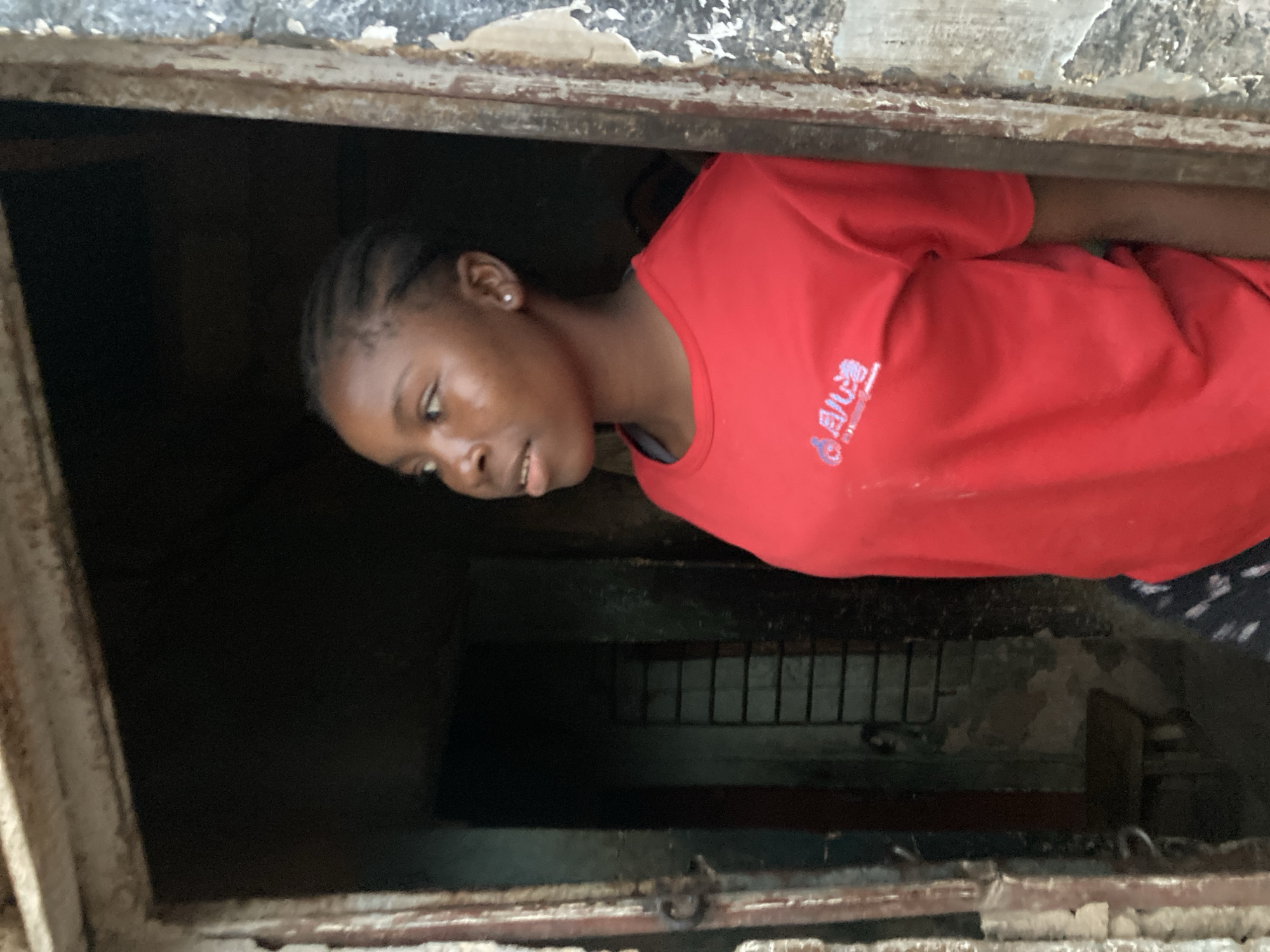 A young Black person, likely a girl, with braided hair is peering out of a dark, enclosed space. She is wearing a bright red t-shirt with some blue text on it. Her head is tilted to her left, and her eyes are looking downwards. The opening she is looking out of appears to be a window or a vent in a rough, textured structure, possibly made of concrete or weathered metal. The background within the opening is dark, but a grated or barred structure is faintly visible. The overall scene is dimly lit, suggesting it might be indoors or a shaded outdoor area. The person's expression is neutral or perhaps curious, as she observes something outside the frame. The text on her shirt is difficult to read clearly but appears to be in an Asian script. The environment looks somewhat neglected or rustic.