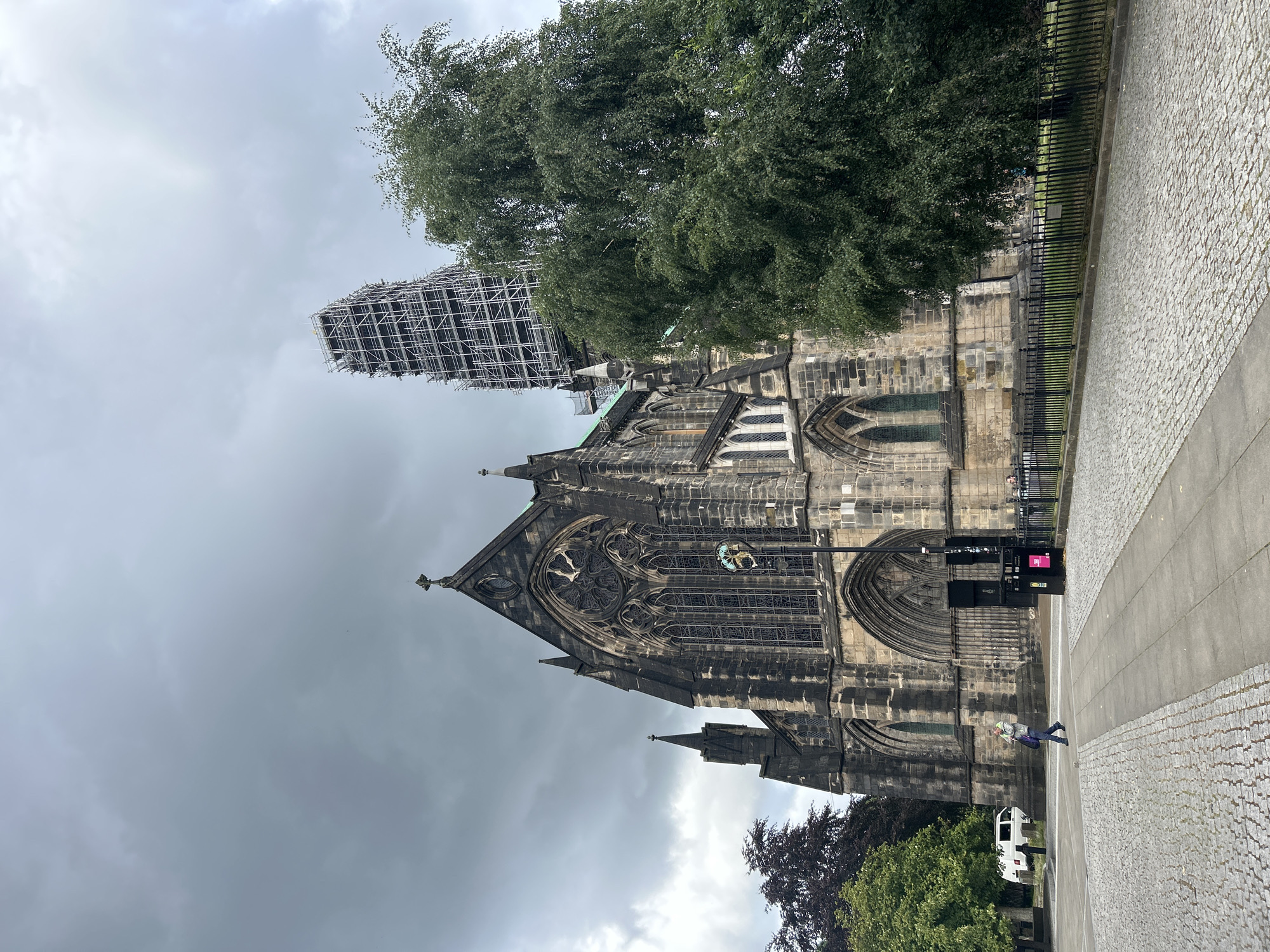 The image shows Glasgow Cathedral, located in Glasgow, United Kingdom, on a cloudy day. The cathedral is a large Gothic structure made of stone, with intricate architectural details including pointed arches, stained glass windows, and spires. Scaffolding is visible on one of the cathedral's towers, indicating ongoing construction or maintenance. In front of the cathedral, there is a paved walkway and a cobblestone area. A large green tree stands to the right of the cathedral, partially obscuring the view of the building. A person is walking on the paved walkway in the foreground, carrying a bag. A white van is parked in the background to the left. A black metal fence lines the area in front of the cathedral. The sky is overcast and grey.