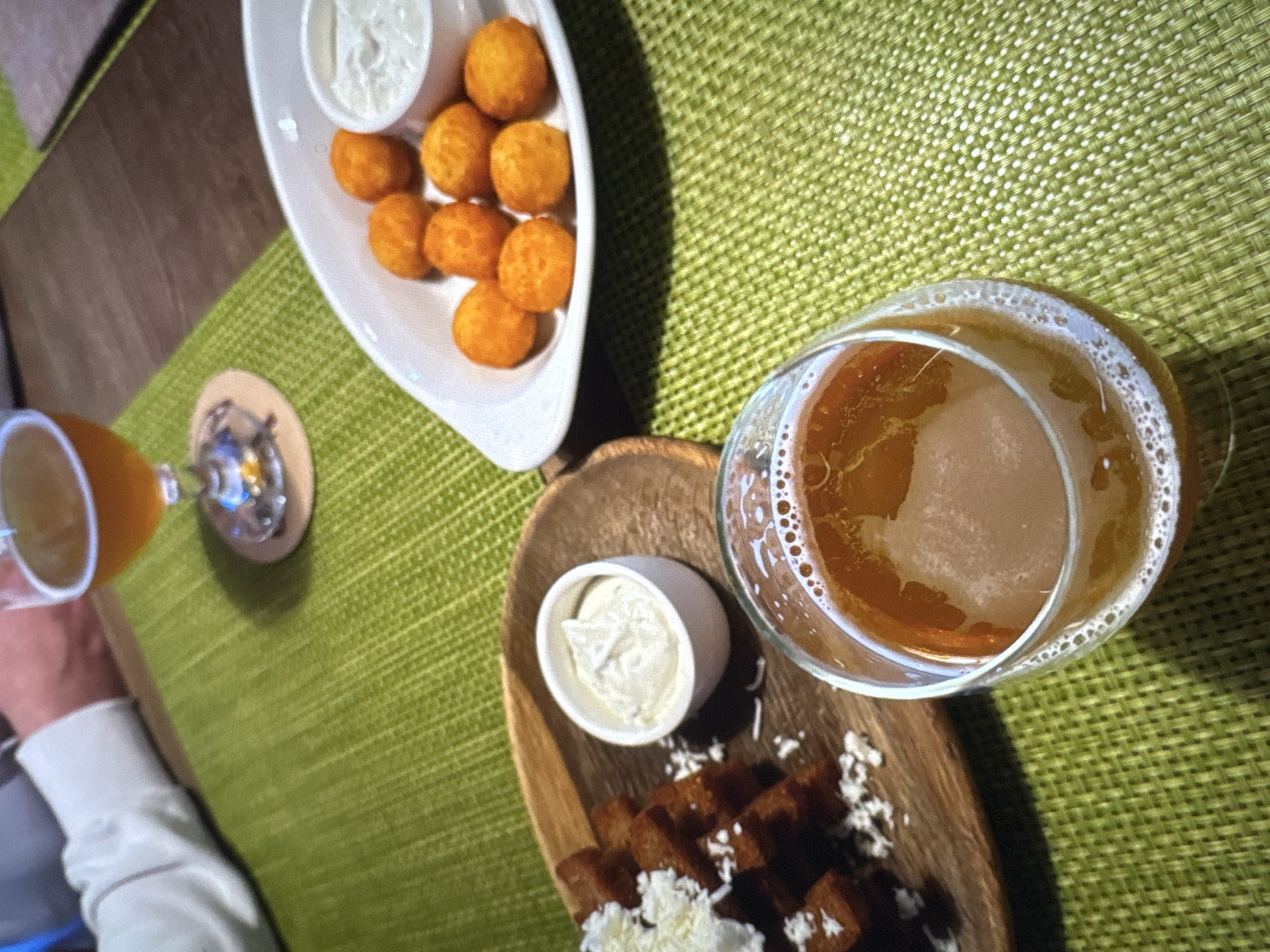 This is a close-up, overhead shot of a table setting with food and drinks. In the foreground, a glass of amber-colored beer with a foamy head sits on a green textured placemat. To the left, a wooden platter holds a pile of dark, bite-sized croutons sprinkled with white grated cheese, next to a small white ramekin filled with a creamy white sauce. In the background, a white serving dish contains several round, golden-orange fried appetizers and a small ramekin of white sauce. Another glass of beer is visible in the upper background, placed on a coaster. A person's arm, wearing a white long-sleeved shirt, is visible on the far left, suggesting a dining or social setting. The overall impression is of a casual meal or appetizer spread. The location is not specified in the image.