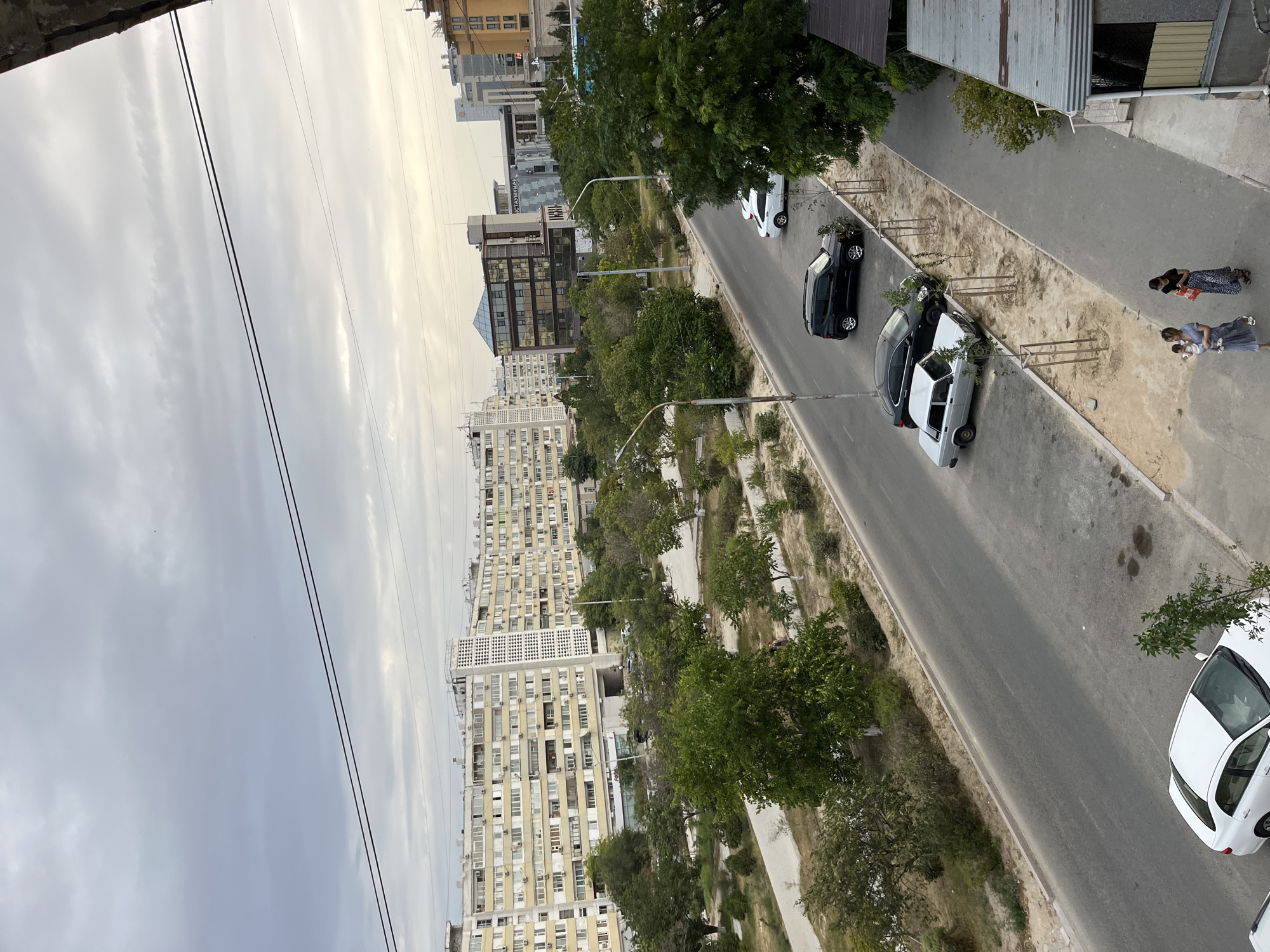 The image depicts a street scene in Aktau, Kazakhstan, with a focus on a wide, paved road running diagonally from the lower left to the upper right. The sky above is overcast with gray clouds. On the left side of the road, there is a wide green space with numerous trees and bushes, and a walking path. Behind this green space, a very long, multi-story apartment building, characteristic of Soviet-era architecture, dominates the background. This building is light-colored and appears to have many balconies and windows.
On the right side of the road, there is a sidewalk lined with young trees planted in sandy soil. Several cars are parked along the street. In the foreground on the sidewalk, two women are walking. One woman is carrying a baby. Both women are dressed in casual clothing. Further down the sidewalk, a building with a modern glass facade and a contrasting darker section is visible, alongside other multi-story buildings, some of which appear to be commercial or office spaces. The overall impression is of a well-established urban environment with a mix of residential and commercial architecture, and a noticeable amount of green space.