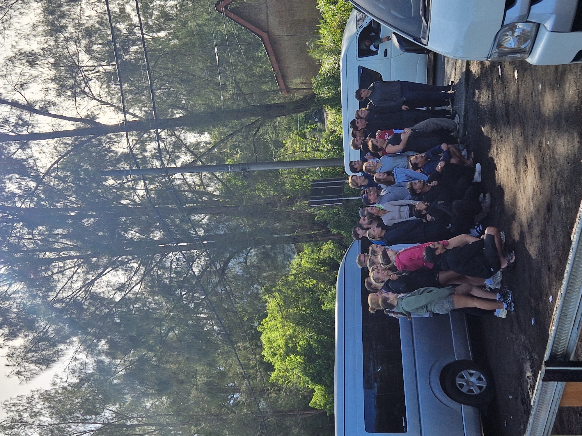In Unknown, Indonesia, a group of approximately 20 people lie on the ground in close proximity, lined up near the side of a road. The group includes both males and females of varying ages, with some wearing athletic clothing. They are all looking upwards. Two white vans are parked nearby, one behind and one to the side of the group. A person is visible inside one of the vans. The environment is wooded, with tall trees lining the roadside. Electrical lines are visible above the group. The ground is dirt and gravel. A metal guardrail is visible on the right side of the frame.