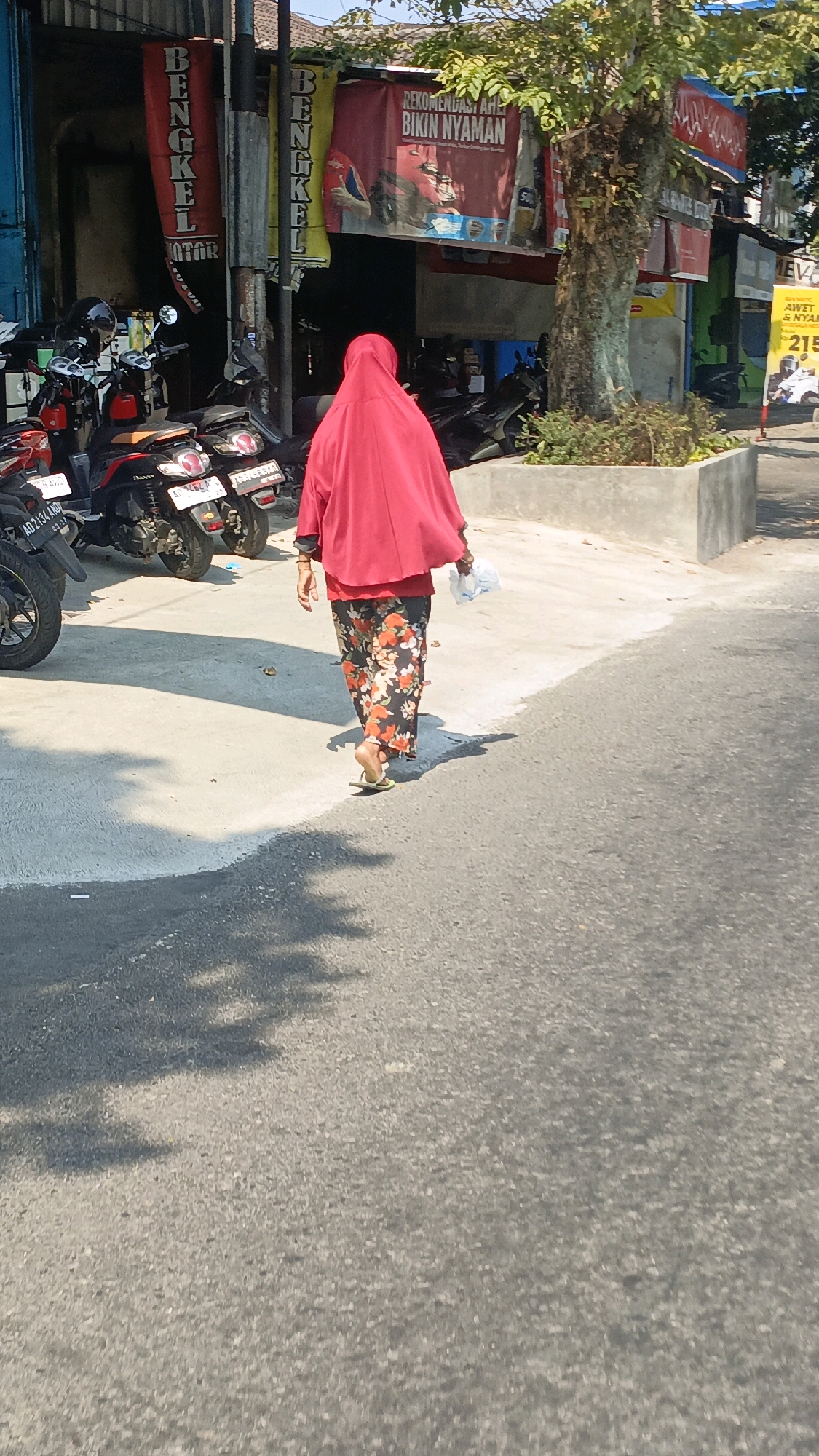 In the city of Sidoharjo, Indonesia, a person wearing a red hijab and floral pants walks away from the camera on a paved road. The person is carrying a white plastic bag in their right hand. To the left of the person, several motorcycles are parked. Behind the motorcycles, there is a building with red and yellow banners that read "BENGKEL MOTOR" (Motorcycle Workshop). Further to the right, a tree stands with some greenery at its base. The ground is a mix of concrete and asphalt, with shadows cast by the tree and a chain-link fence. The overall scene appears to be a street in a commercial area.
