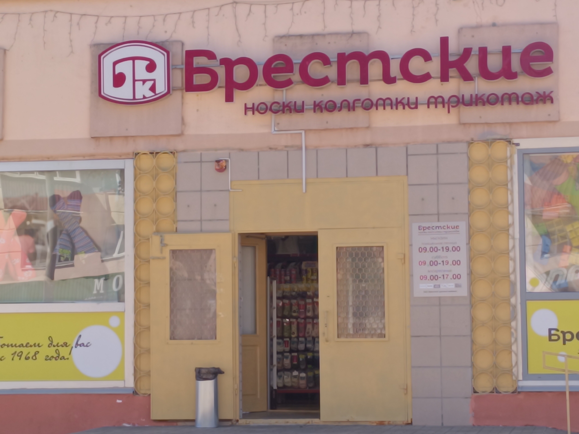 This is a photograph of a store named "Брестские" (Brestskiye) located in the city of Orsha, Belarus. The store specializes in socks, tights, and knitwear, as indicated by the signage. The storefront features a large red and white logo with the Cyrillic letters "ТК" inside a rounded rectangle. Above the entrance, the store name is displayed in large, magenta-colored Cyrillic letters, with the tagline "носки колготки трикотаж" (socks, tights, knitwear) underneath.
The entrance to the store is a double door, with the left door ajar, revealing shelves stocked with various items. To the left of the entrance, a large display window showcases clothing items, including colorful socks and what appear to be knitted garments. A yellow sign with white lettering and circles is visible on the lower part of this window, which seems to advertise the store's long history, possibly stating "Работаем для вас с 1968 года!" (We have been working for you since 1968!).
To the right of the entrance, a smaller sign displays the store's operating hours: 09.00-19.00 for weekdays and Saturdays, and 09.00-17.00 for Sundays. The building's facade is a light beige color, with decorative vertical yellow cylindrical elements flanking the entrance and parts of the windows. A silver cylindrical trash can is placed to the left of the entrance. The overall scene depicts a commercial establishment in an urban setting. There are no people visible in the photograph.