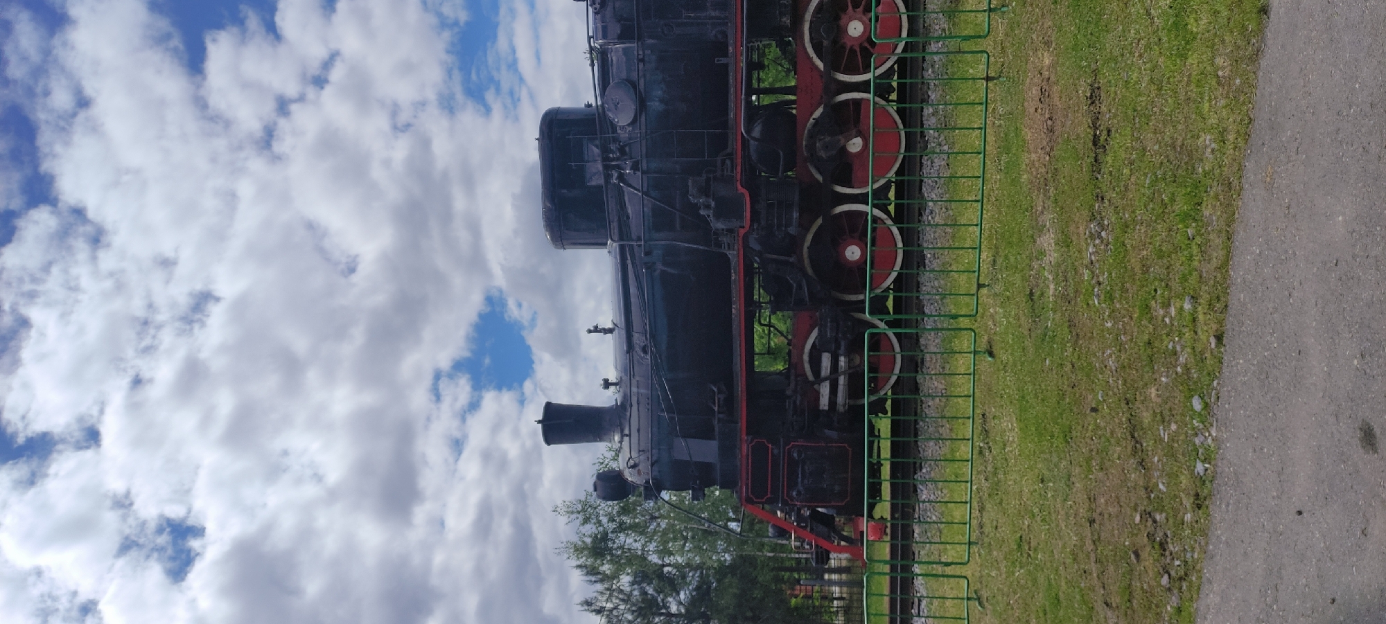 The image captures a black steam locomotive, likely a preserved historical piece, resting near a green fence and a patch of green grass. The background shows a partially cloudy sky. The location is Ivanovskoe, Russia, based on the broader context. The train's red wheels and detailing contrast against its black body. A paved path is visible alongside the grass. Trees can be seen near the base of the locomotive.