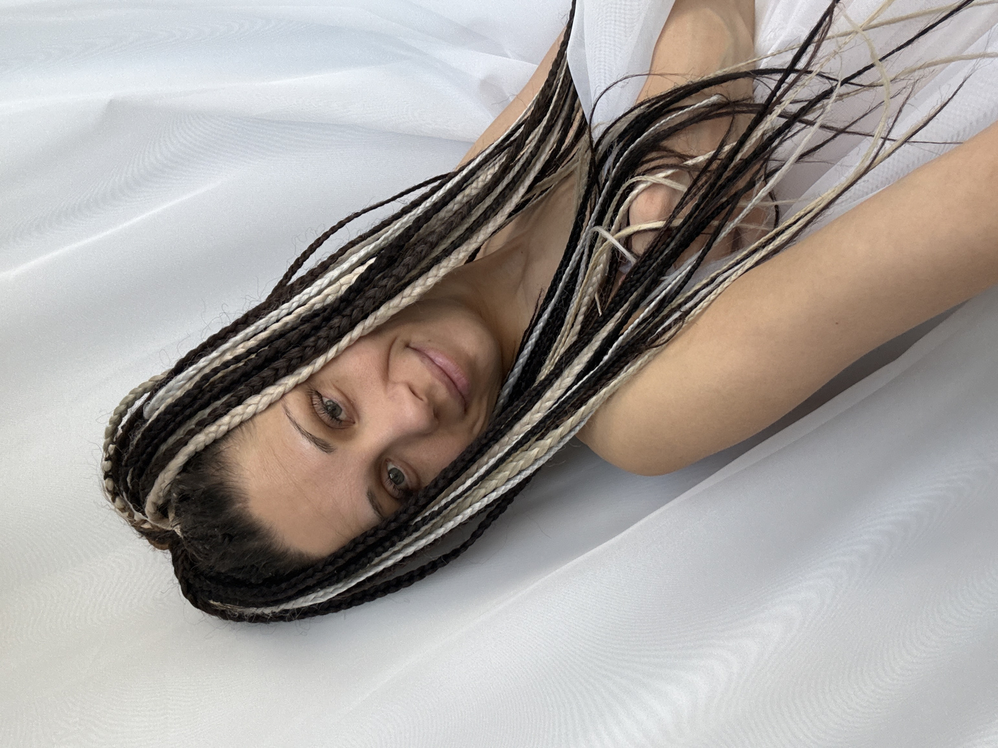 A woman with black and white braided hair is lying on a white surface. Her arms are raised and bent, partially obscuring her face. She is looking directly at the camera with a slight smile. The background is a white, draped fabric. The location is unknown.