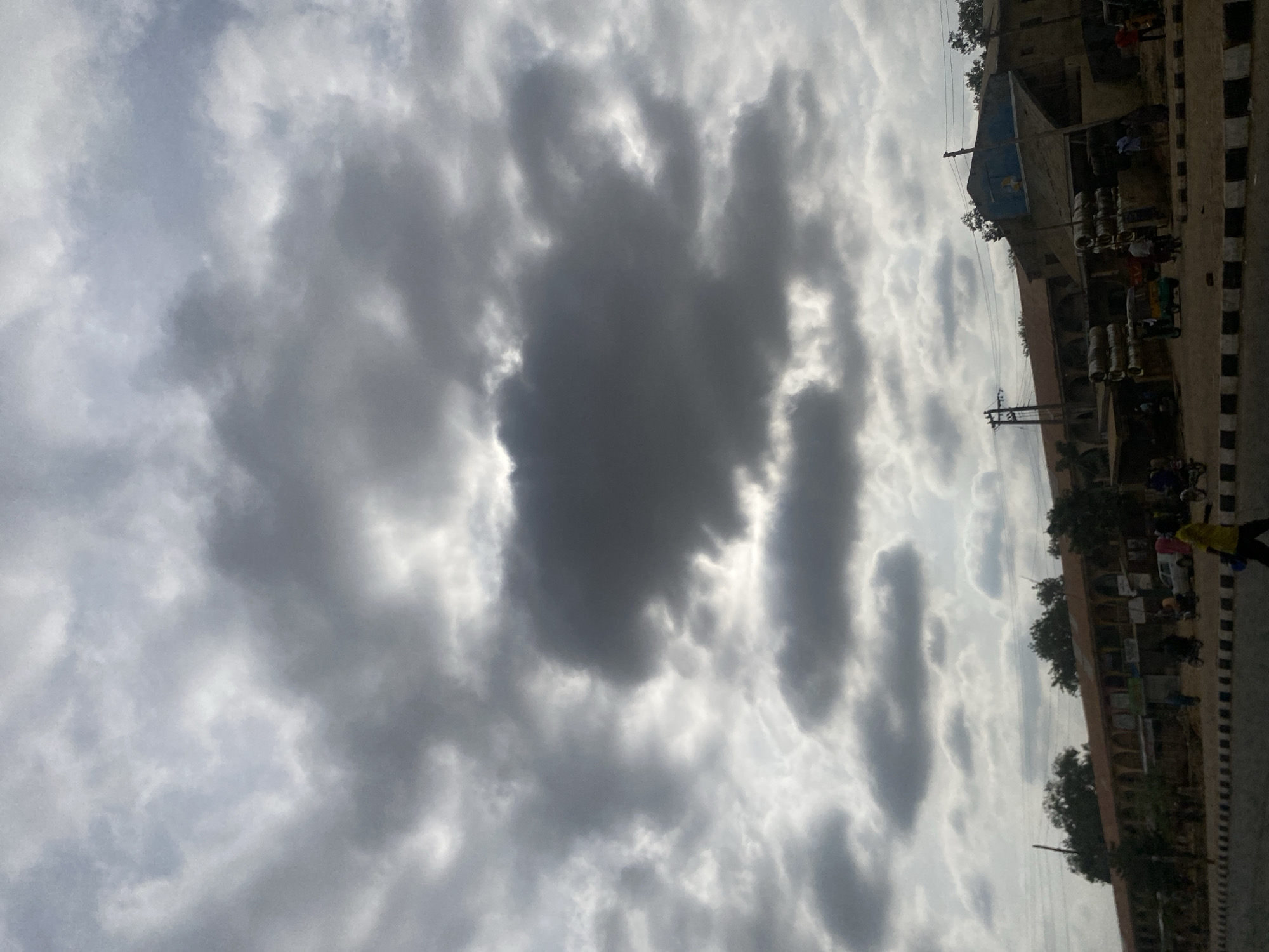 The image captures a cloudy sky over Tudun Wada, Nigeria. A mix of dark gray and light gray clouds fills most of the frame. Along the right edge of the image, there is a street with buildings, trees, people, and parked vehicles visible. There is a sidewalk with a black-and-white painted curb. The sky is overcast.