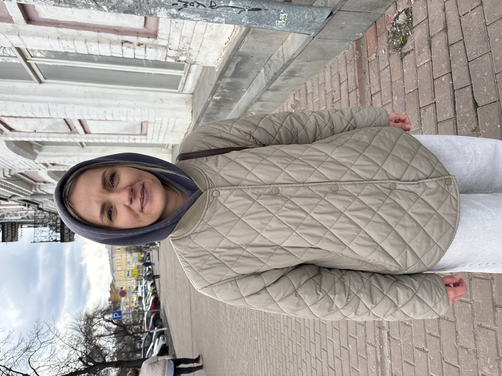 A woman in a quilted jacket and gray hoodie stands on a brick sidewalk in Ulyanovsk, Russia. The sidewalk borders a building with white window frames and a metal pipe. In the background, there is a street with parked cars, signage, and additional buildings. The sky is overcast.