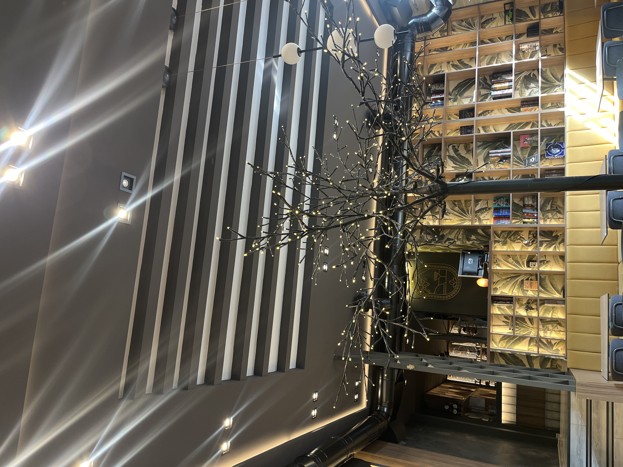 This image depicts the interior of a modern cafe or restaurant located in Plovdiv, Bulgaria. The scene is characterized by a high ceiling with a dramatic lighting design featuring exposed beams and recessed lights that cast strong, diagonal beams of light across the space. A prominent feature is a decorative tree sculpture with numerous small, illuminated lights, positioned in the foreground and partially obscuring the view of the background.
The background reveals a stylized interior design. A long, cushioned yellow bench runs along the wall, above which is a large, grid-like shelving unit filled with books and decorative objects. The wall behind the shelves is covered in a tropical-themed wallpaper featuring large green leaves on a yellow background. To the left of the shelving unit, there is a counter area with a computer monitor and a decorative lamp. Further left, a staircase leads to an upper level, with the interior walls adorned with geometric patterns and wood paneling. The overall ambiance suggests a contemporary and chic establishment. There are no people visible in the image.