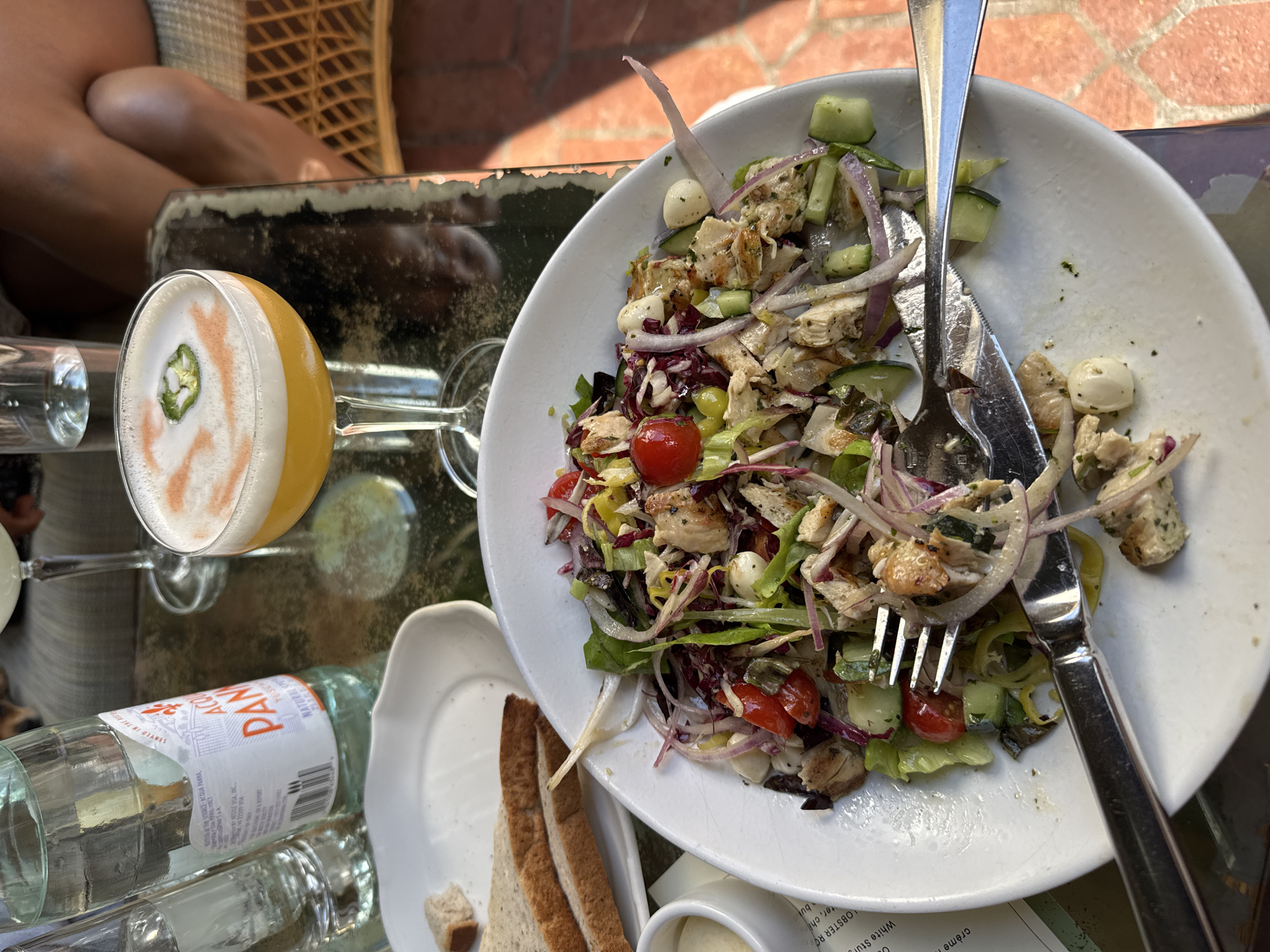 Here is a detailed description of the image:
**Scene:** The image captures a meal scene, likely at an outdoor dining area.
**Location:** While not explicitly stated, the OCR text on the water bottle suggests a location serving Acqua Panna water. The text mentions "NESTLE USA, INC," and "SERVED IN THE BEST," alluding to a restaurant or establishment in the United States. Although the location of the photograph is mentioned as West Hollywood, United States of America, the details were not visible in the photograph.
**Subjects:**
* A person's legs are visible in the background.
* The primary focus is on a half-eaten salad on a white plate.
**Objects:**
* **Food & Beverages:**
* A mostly-consumed salad with ingredients like lettuce, tomatoes, cucumbers, red onions, and what seems to be grilled chicken or fish. Small mozzarella balls are also present.
* A cocktail in a stemmed glass, topped with foam and a slice of jalapeño.
* A bottle of Acqua Panna water.
* A small dish of some kind of sauce
* Sliced bread on a small plate.
* **Tableware:**
* A white plate with the salad on it.
* Fork and knife resting on the plate.
* Several drinking glasses.
* A small white plate for the bread.
* **Surfaces:**
* A table with a reflective surface, possibly glass or a polished material.
* A wicker chair in the background, partially visible.
* **Miscellaneous:**
* A printed menu or leaflet with text on the lower right side.
**Actions & Interactions:**
* Someone has been eating the salad, as evidenced by its partly consumed state and the cutlery.
* A drink has been consumed or is about to be.
**Environment:**
* The overall environment appears to be bright and sunny, likely outdoors or in a well-lit area.
* The table setup suggests a casual or fine-dining setting.